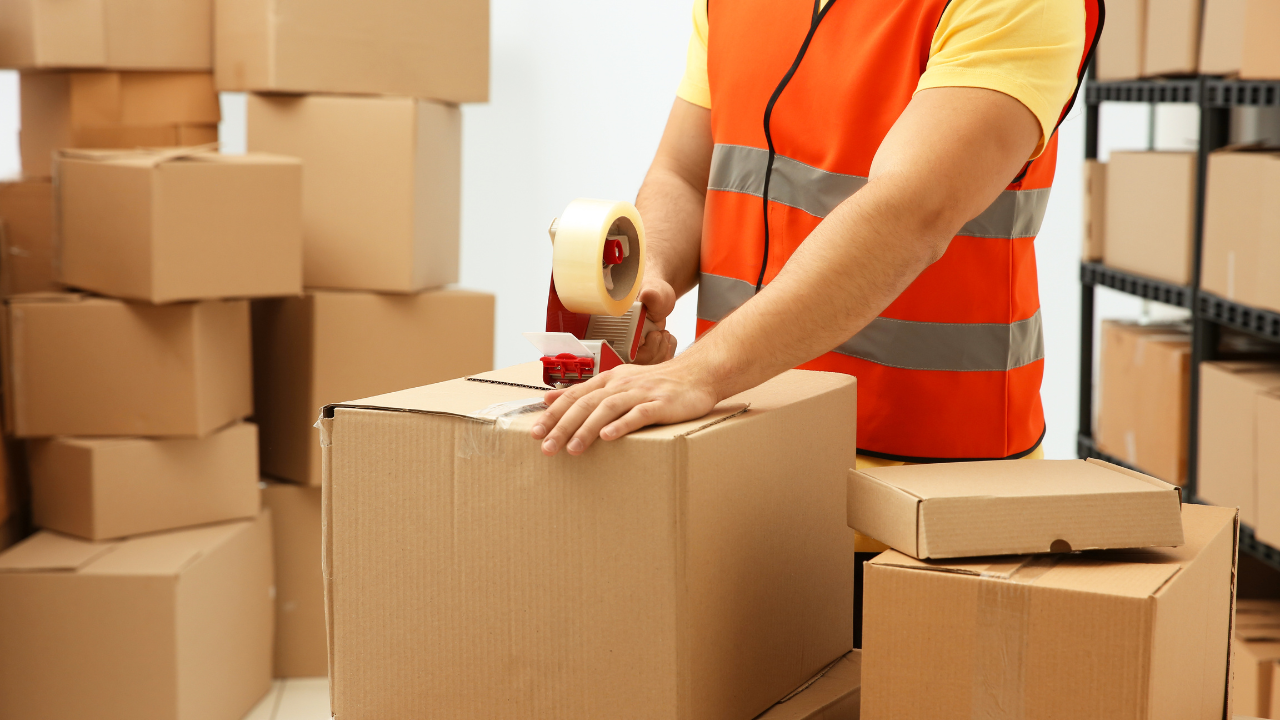 Person in orange vest taping a cardboard box in a warehouse setting, surrounded by stacked boxes.