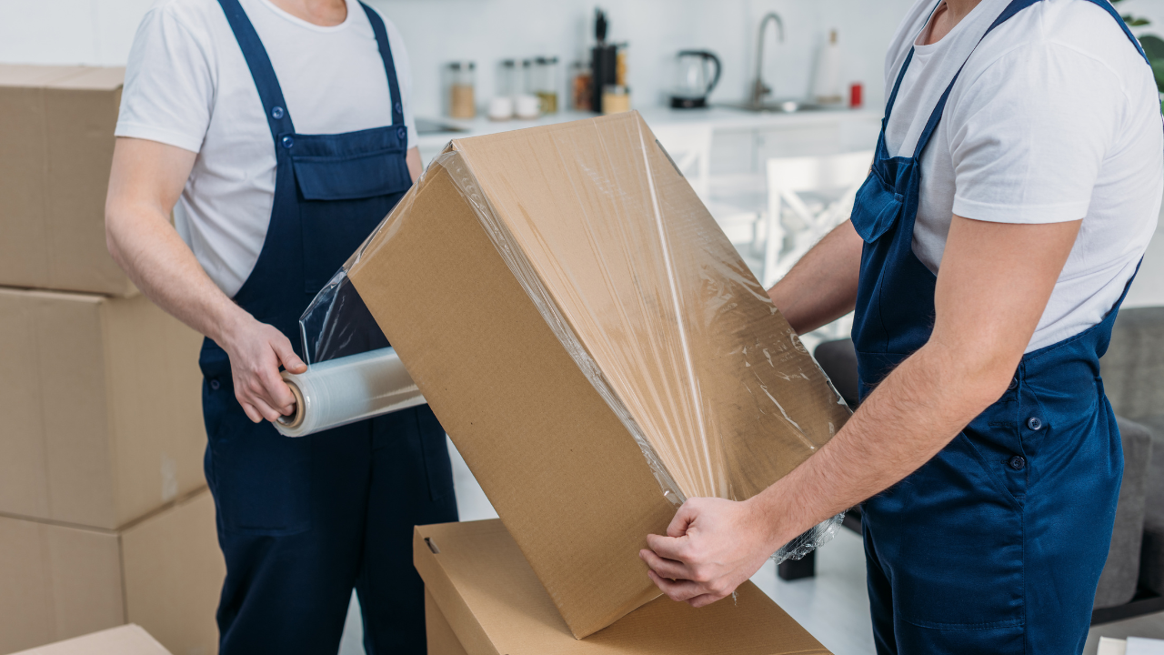 Two movers wrapping a cardboard box with clear plastic wrap in a home setting.