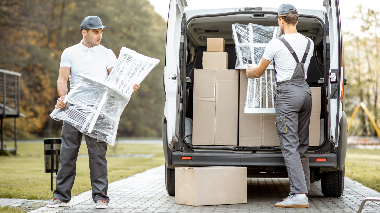 Two movers load a van with wrapped furniture and boxes outdoors.