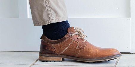A person's leg with a brown leather shoe, blue sock, and tan pants against a white door.