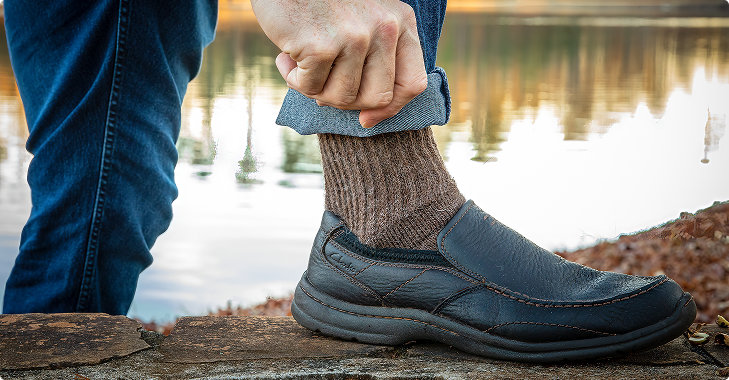 Person cuffing blue jeans, revealing brown sock and black slip-on shoe. Outdoors, near water.
