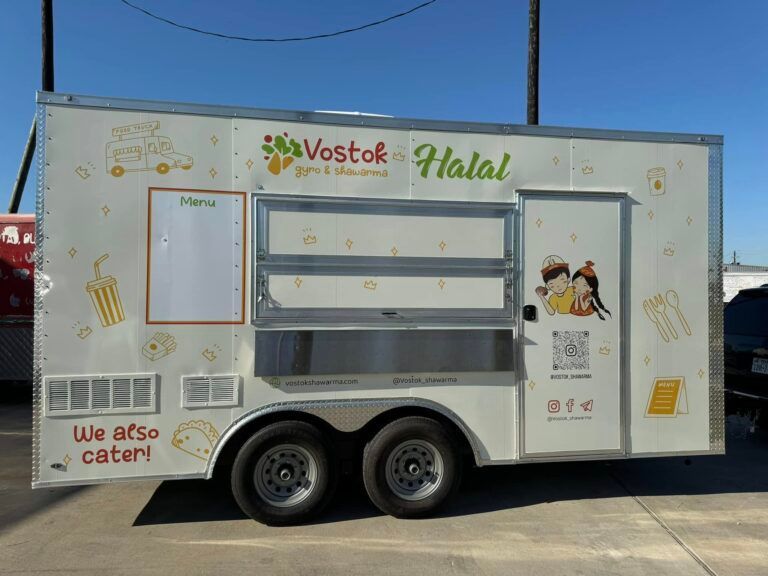 A white Vostok Halal food trailer parked outdoors, featuring a menu board, service window, and catering information.