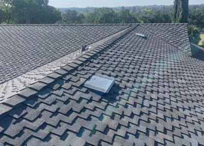 Gray asphalt shingle roof with a skylight, vent, and trees in the background on a sunny day.