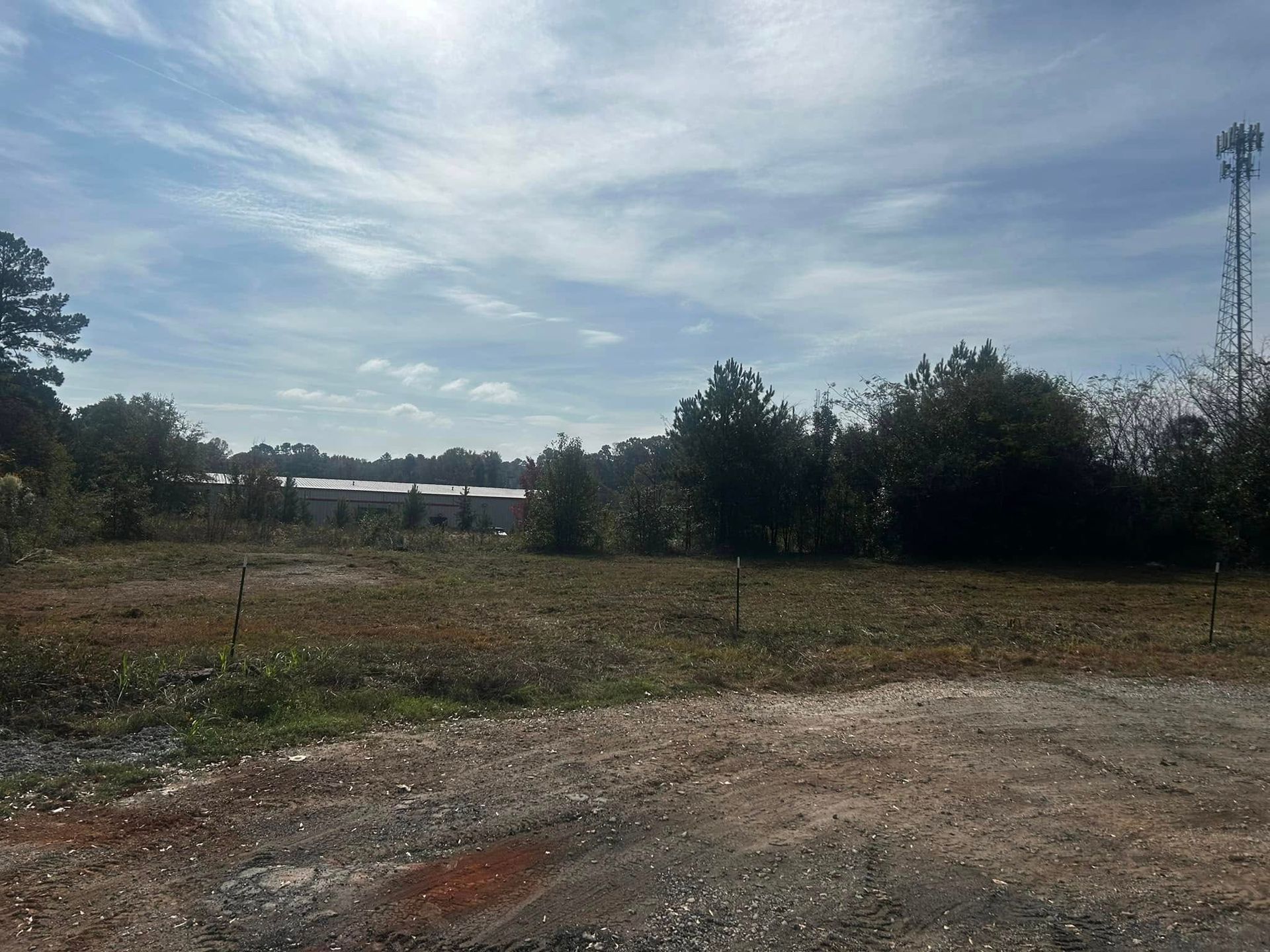 Open lot with gravel foreground, dry grass, trees, a building, and cell tower under a cloudy sky.