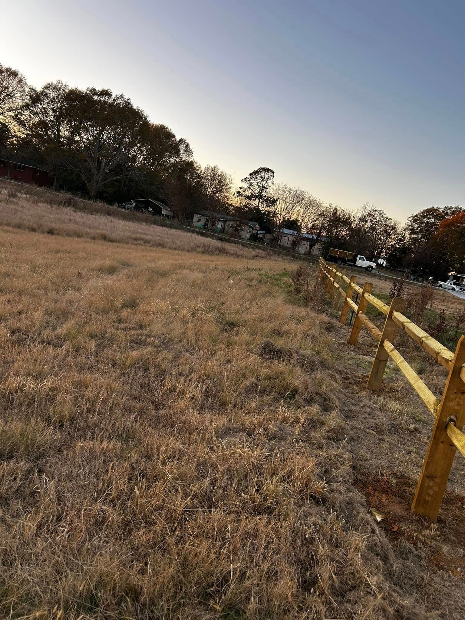 A golden-hued field with a split-rail fence, trees, and buildings against a clear sky.