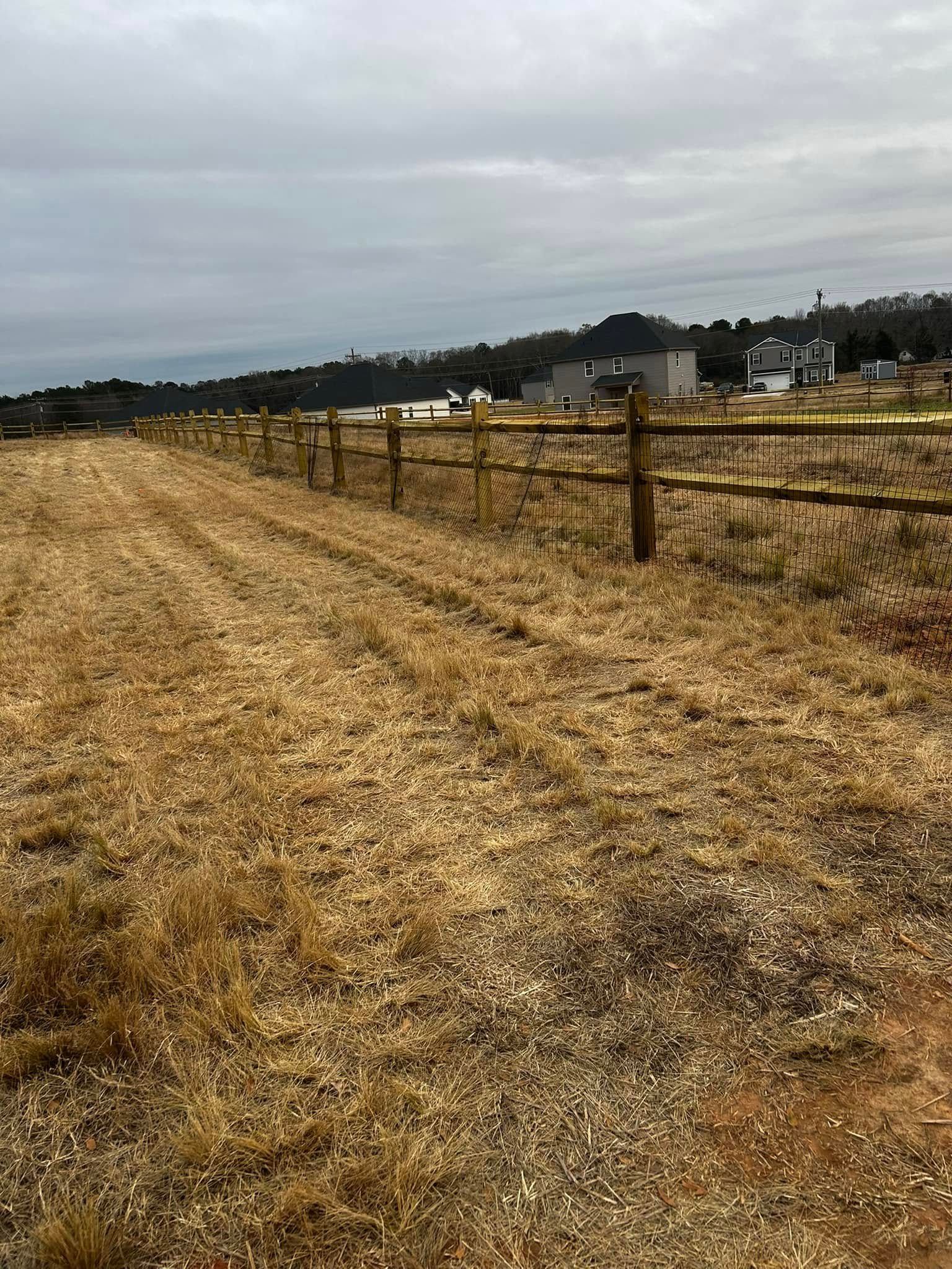 A dirt path runs between a wooden fence and dry grass, with houses in the distance under a cloudy sky.