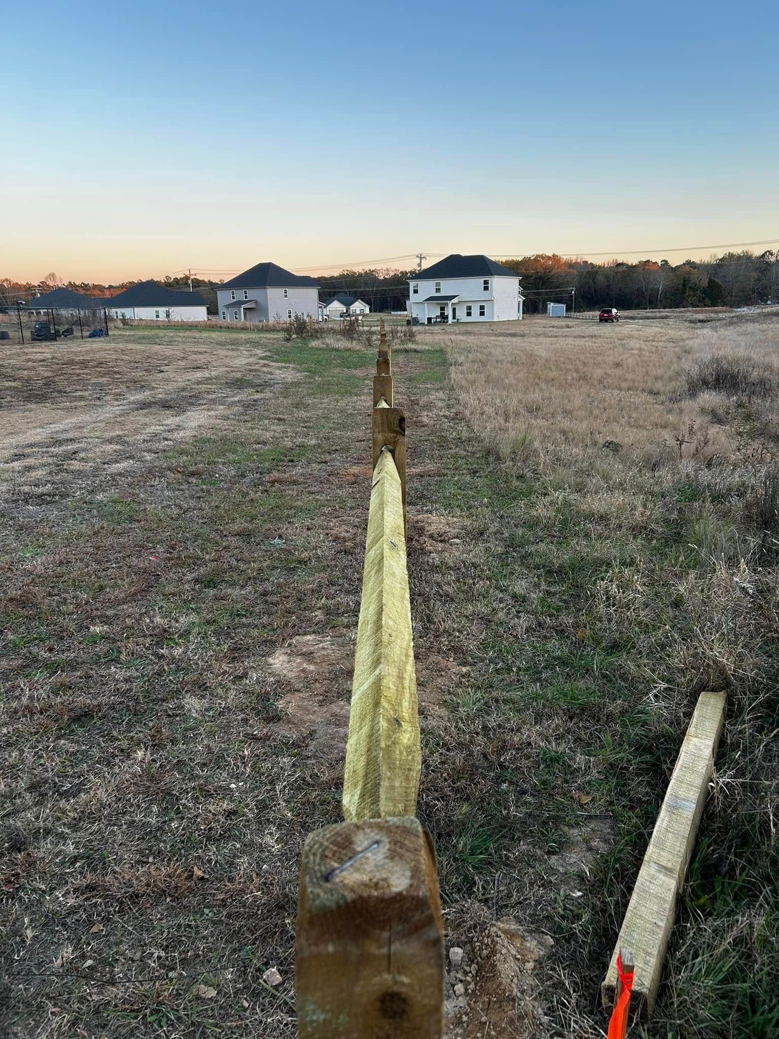Posts for a fence line in a grassy field with houses in the background at dusk.
