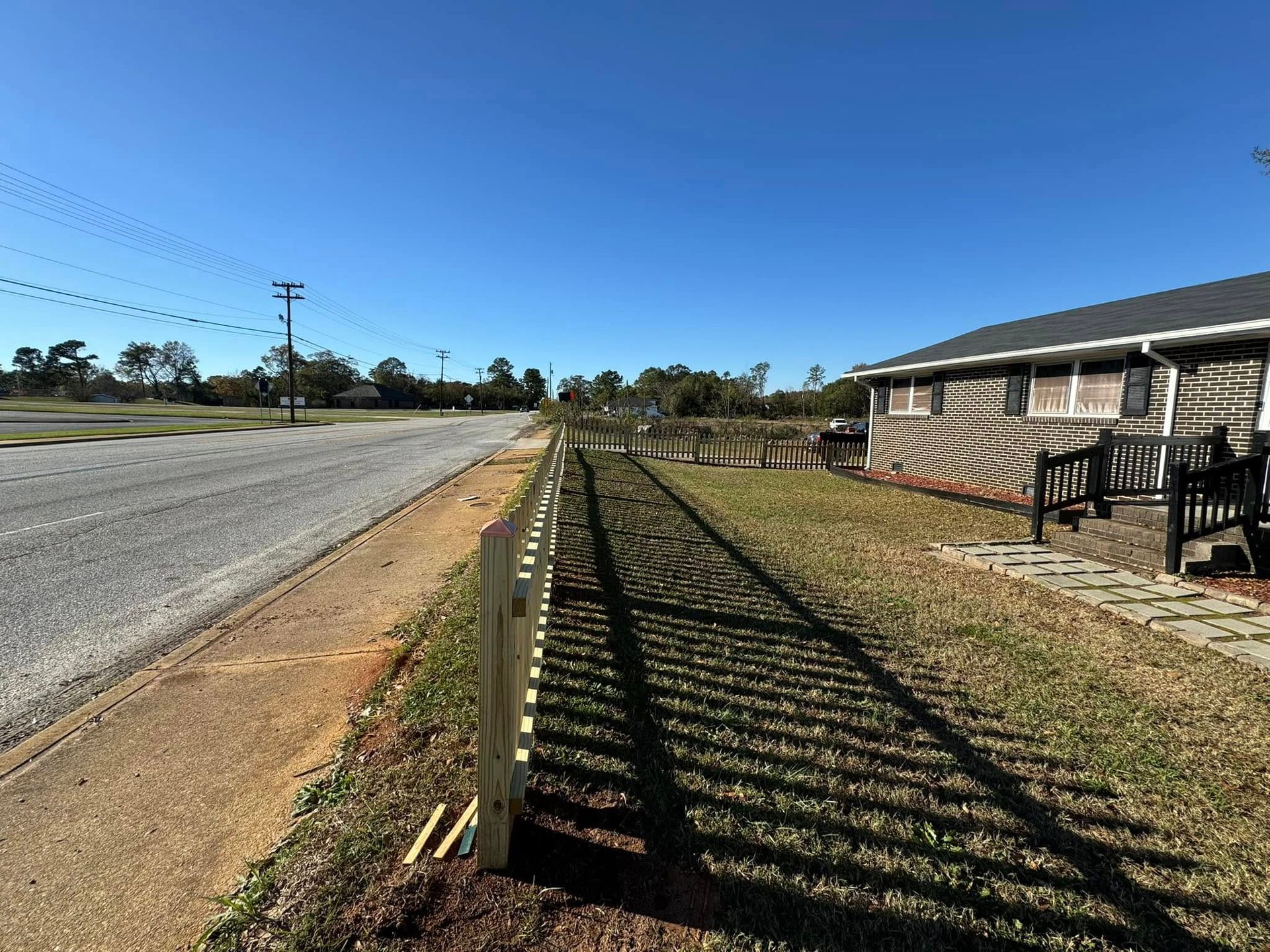 A white picket fence borders a grass lawn next to a road, with a house on the right under a clear blue sky.