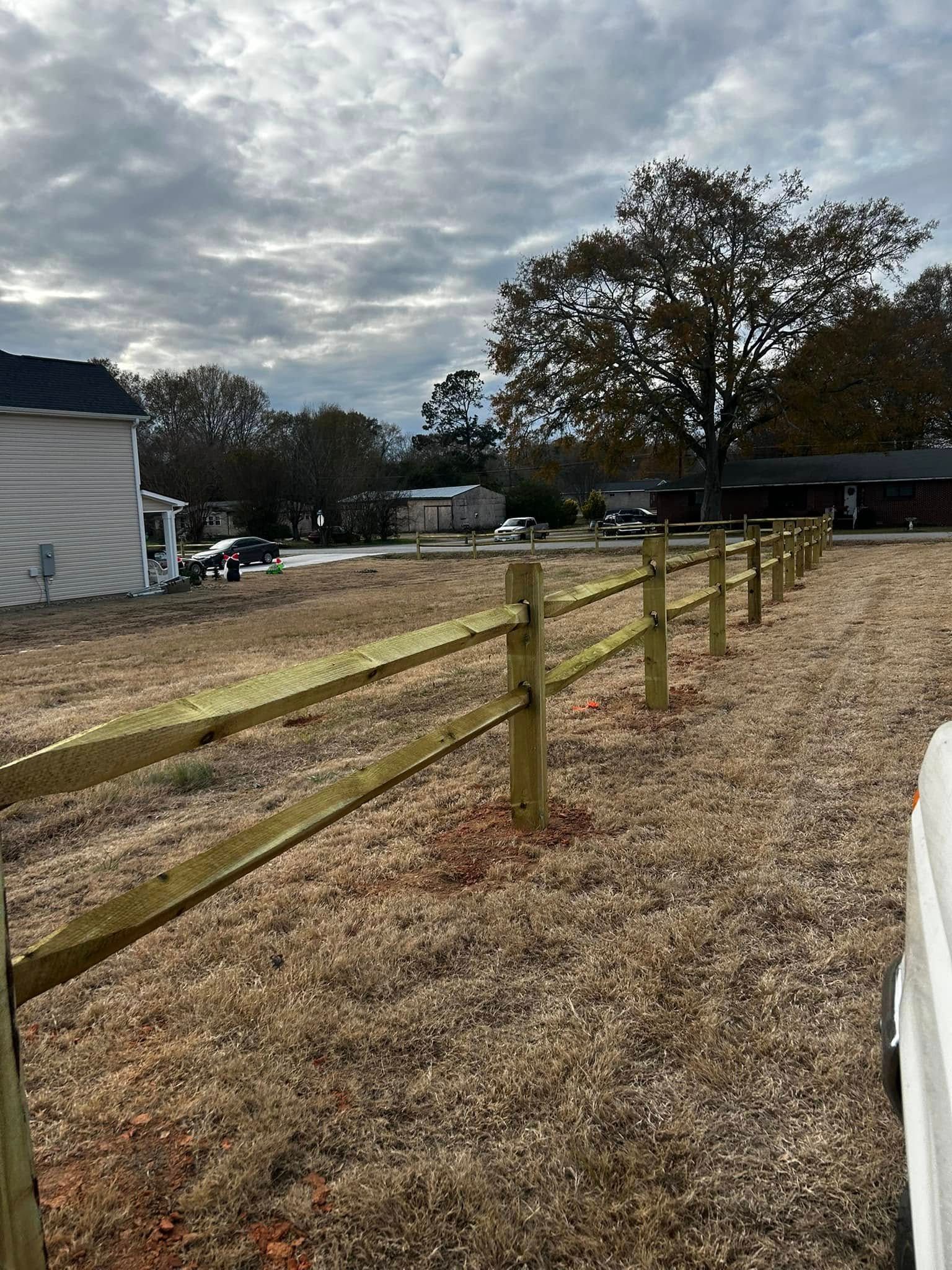 Wooden two-rail fence along a grassy yard, under a cloudy sky, with houses in the background.