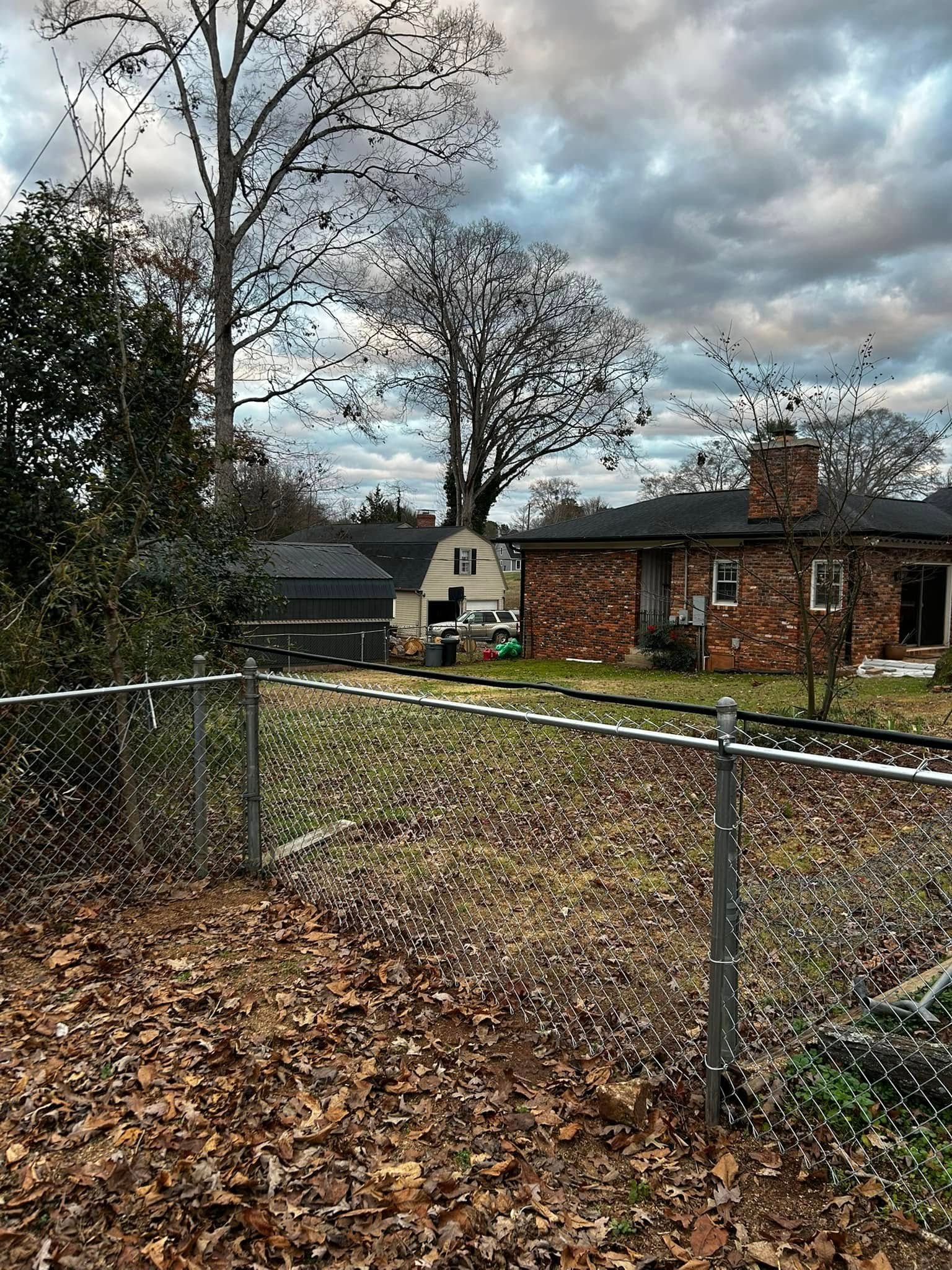 Chain-link fence in front of a yard with dried leaves. A brick house and shed are in the background. Overcast sky.