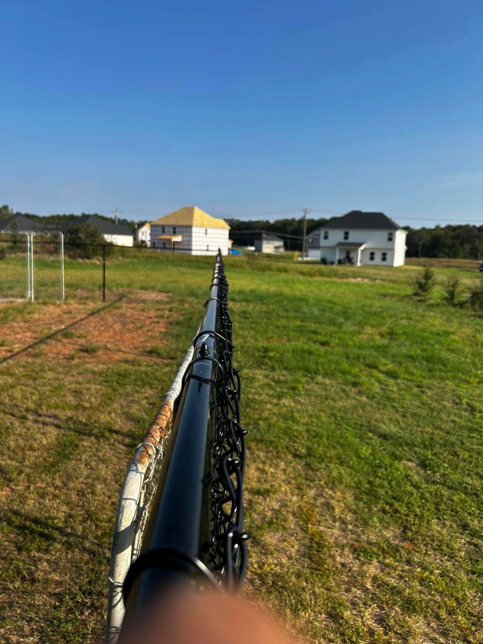 Black fence in a grassy field with houses in the background under a blue sky.