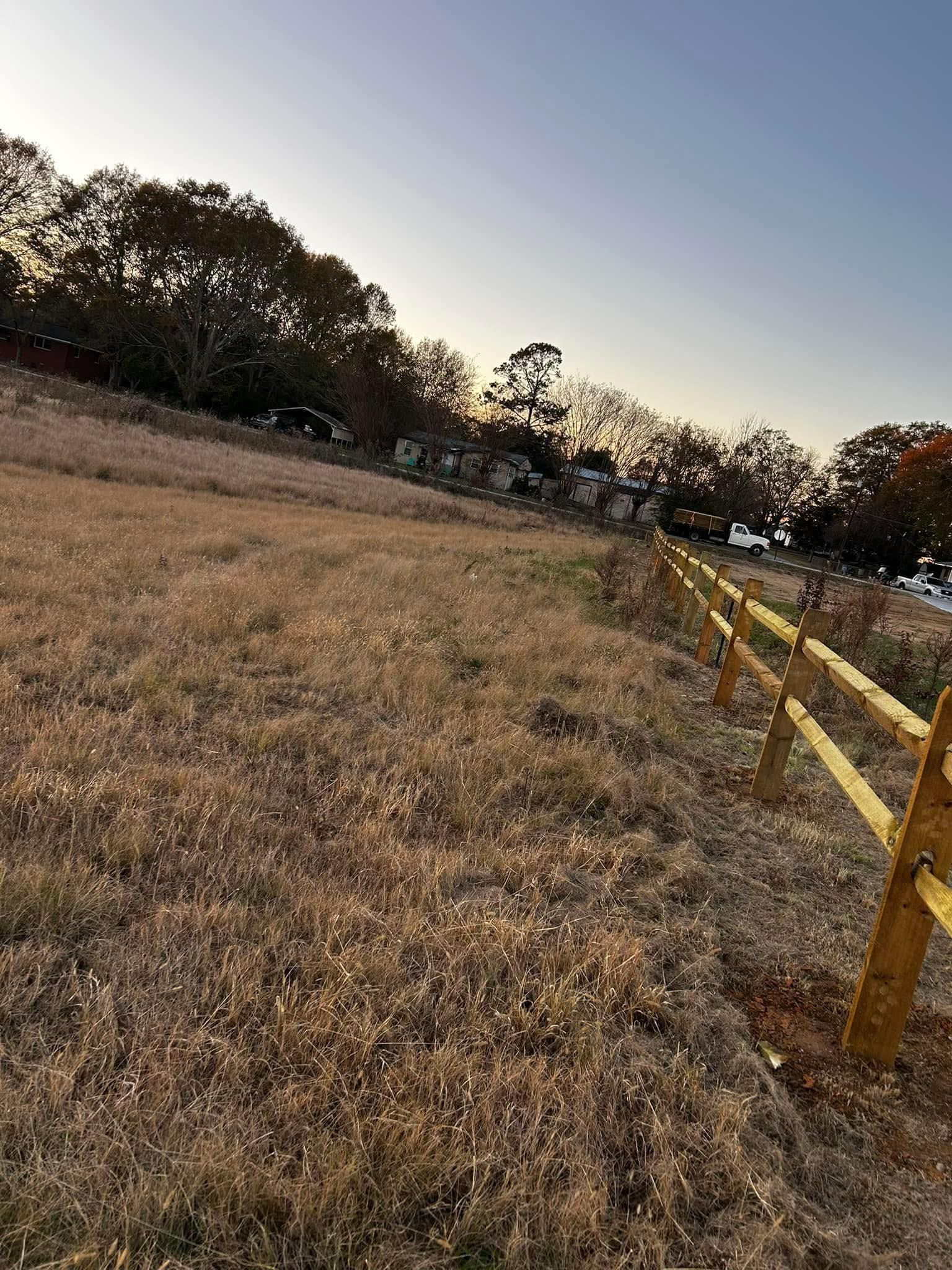 A golden-lit field with dry grass and a wooden fence, with trees and a light-blue sky in the background.