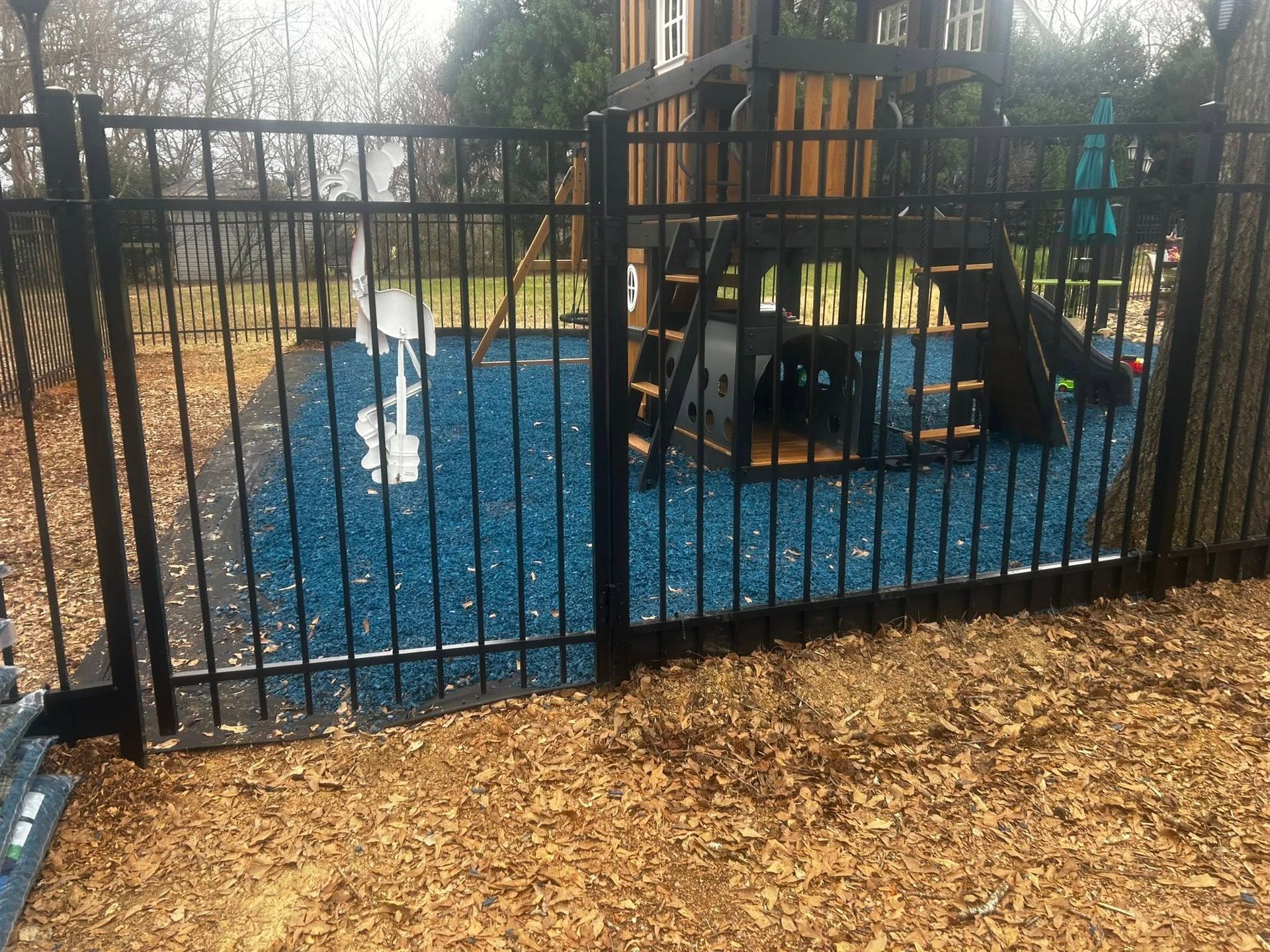 Black fence encloses a playground with blue rubber surface and wooden play structure.
