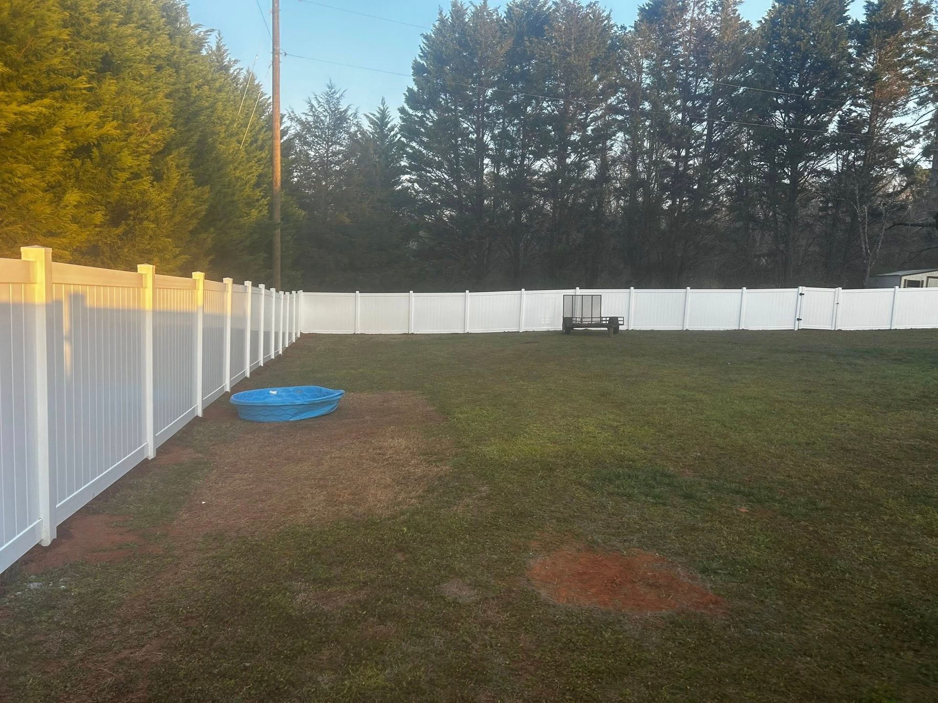 A white fenced backyard with a small blue pool on the lawn. Trees and blue sky in the background.
