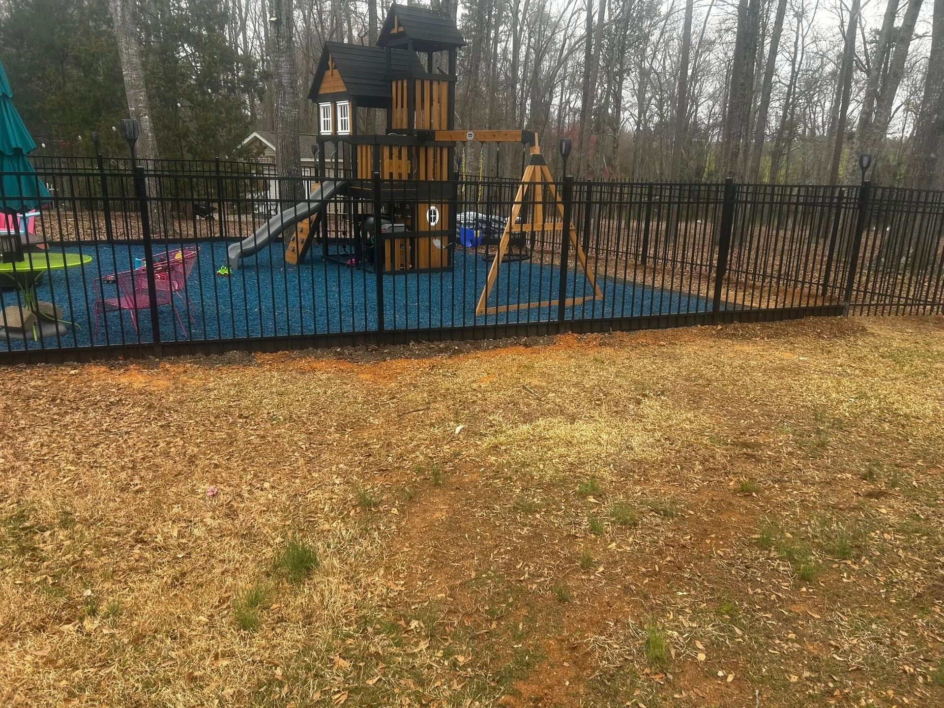 Black fence encloses a playground with a wooden play structure and blue rubber surface; dry grass in foreground.