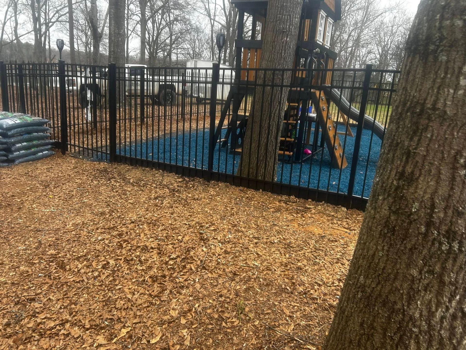 Black fence surrounds playground with blue ground cover, trees, and mulch.