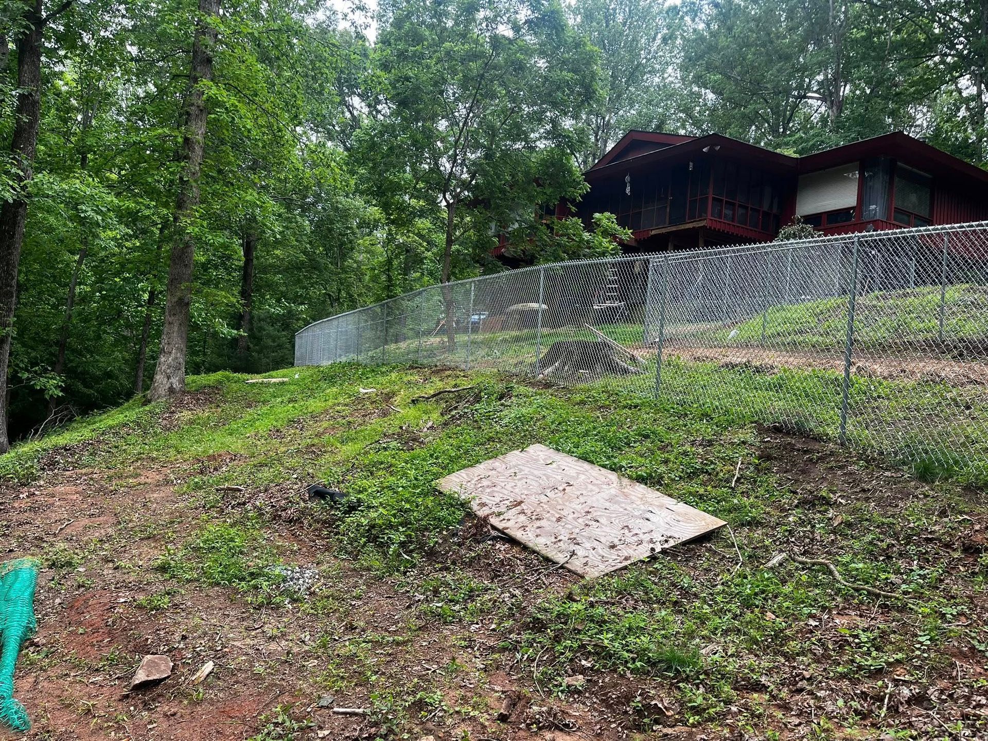 A house sits behind a chain-link fence on a grassy hill, trees in the background.
