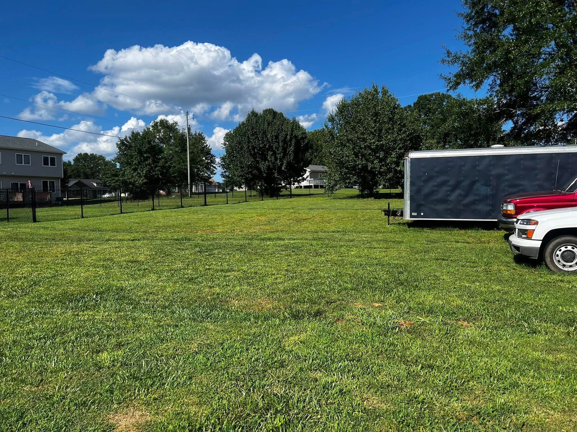 Grassy lot with trees, blue sky, trailer, and a red and white truck. A building and fence are in the distance.
