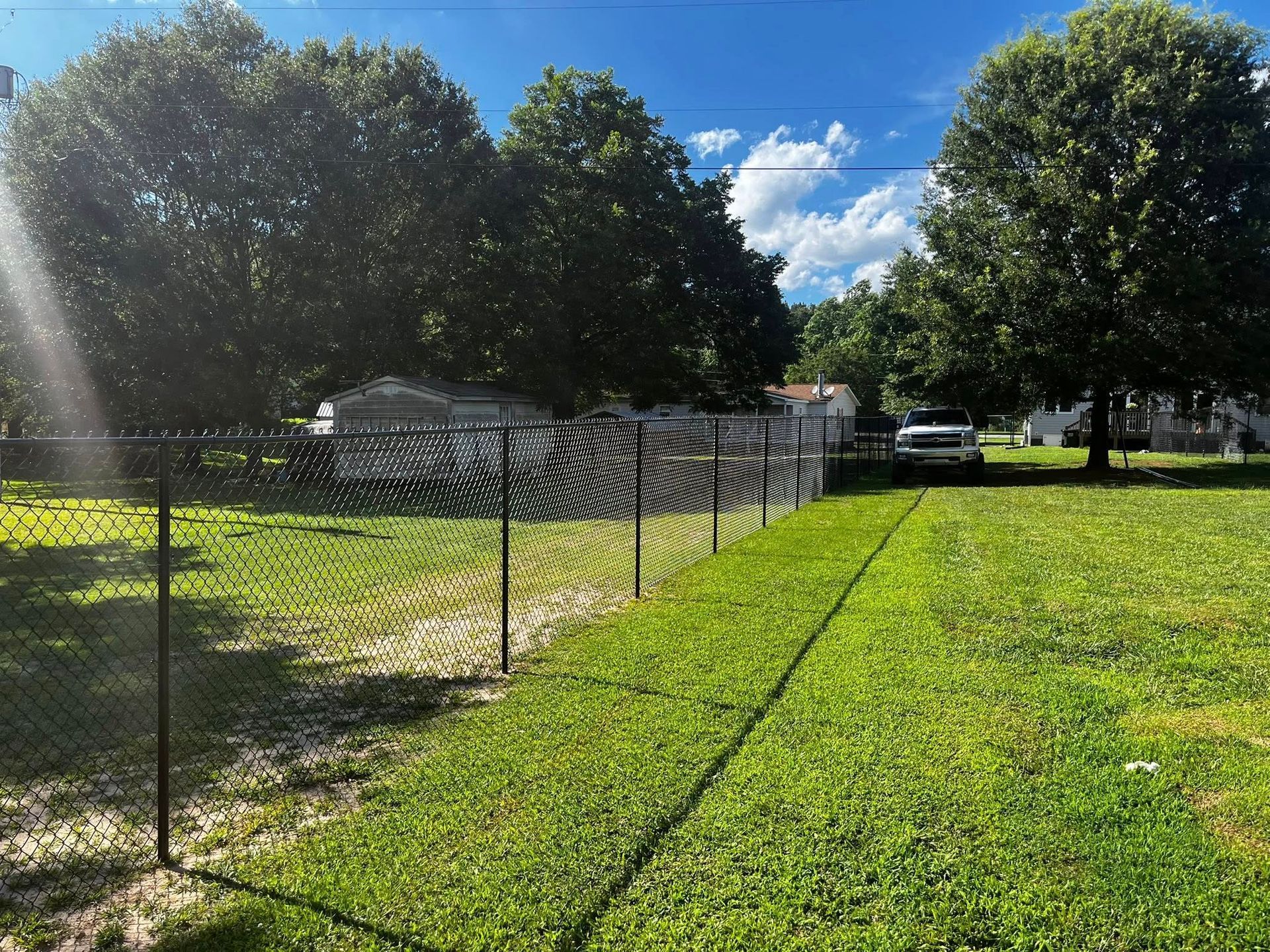A black chain-link fence divides a grassy yard. A car is parked ahead on grass. Trees and sky are in background.