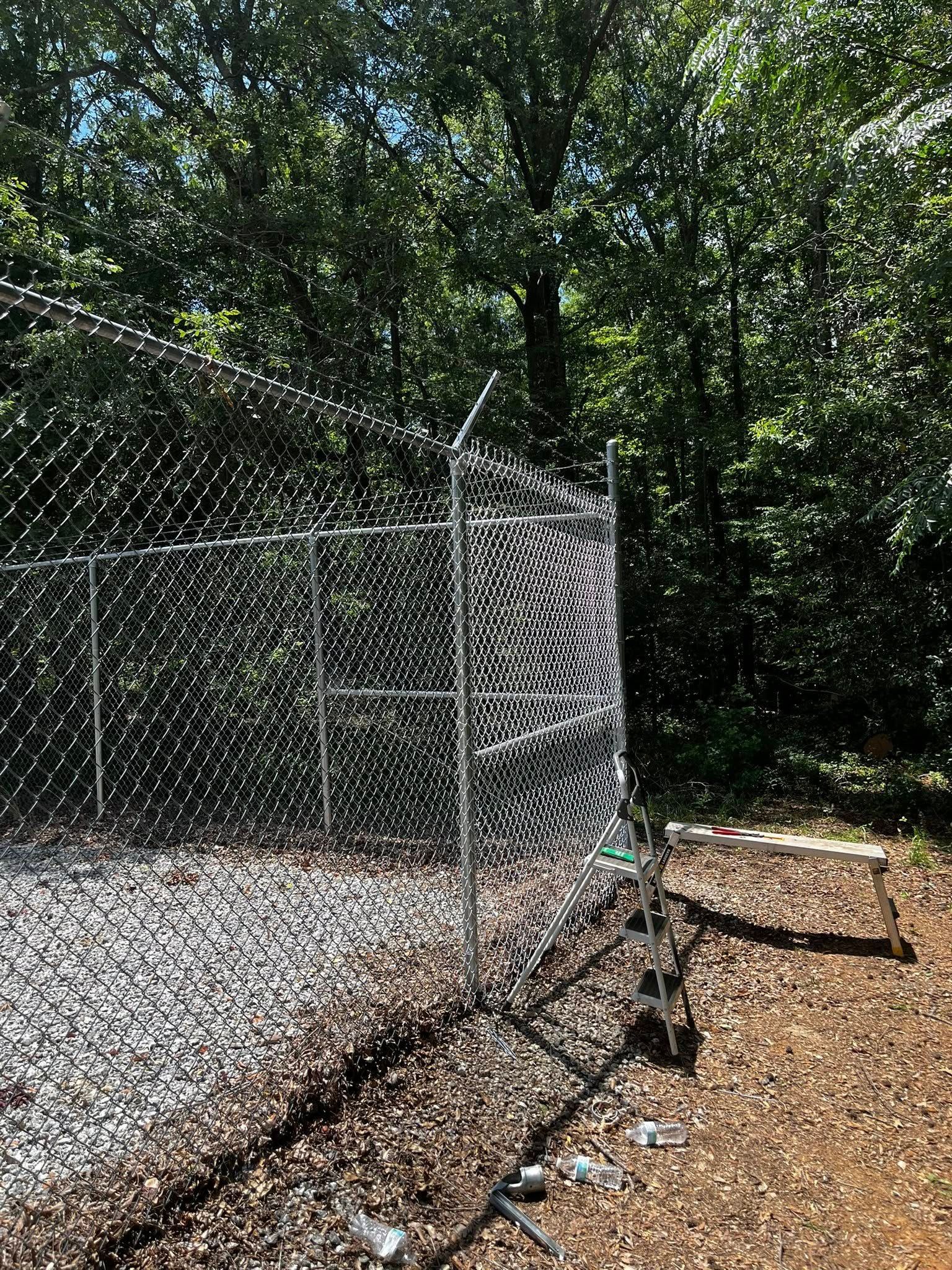 Chain link fence in a forest, with razor wire at the top. Ground covered in gravel and leaves.