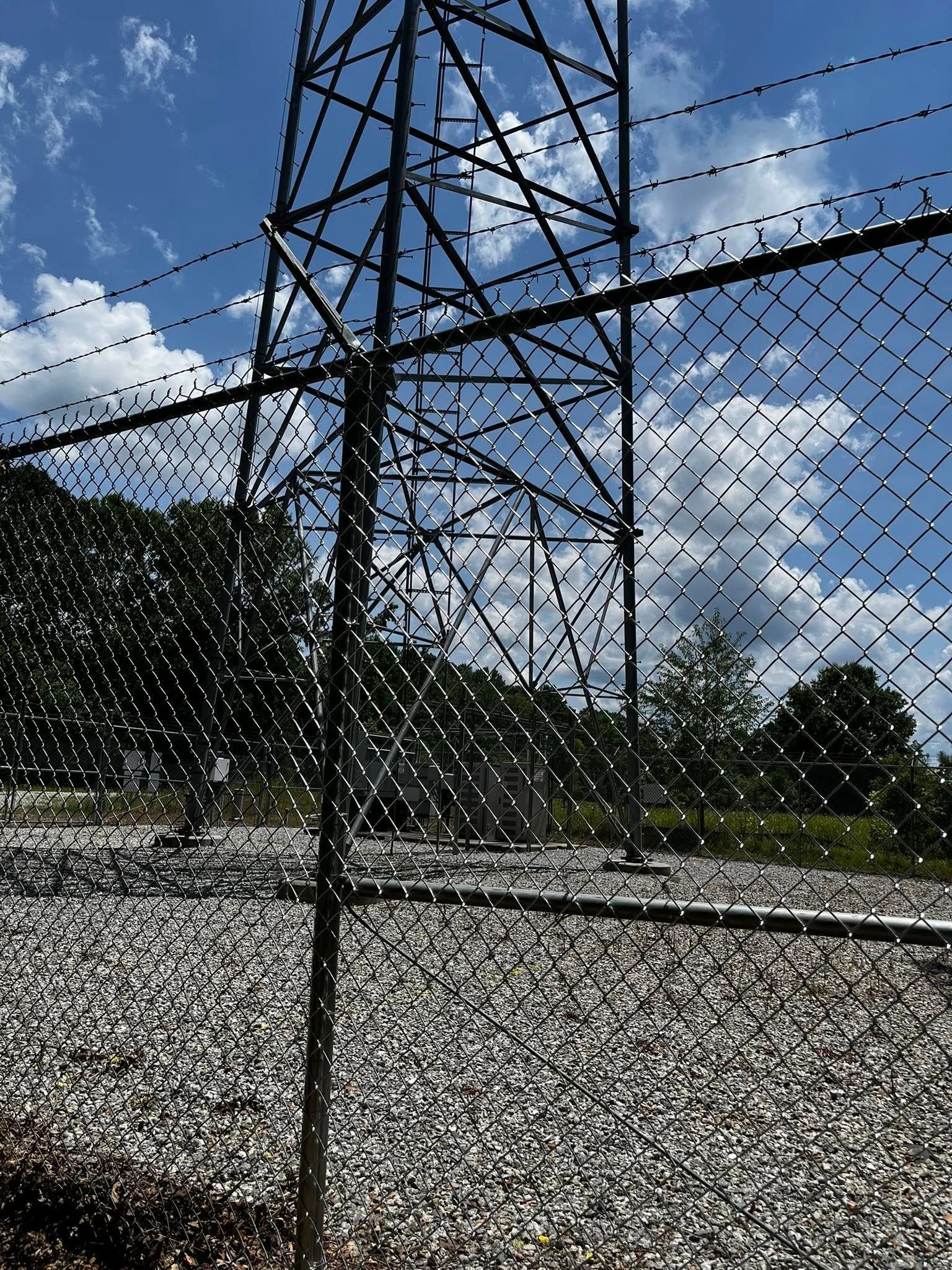 A tall electrical tower behind a chain link fence and gravel, blue sky, and trees in the background.
