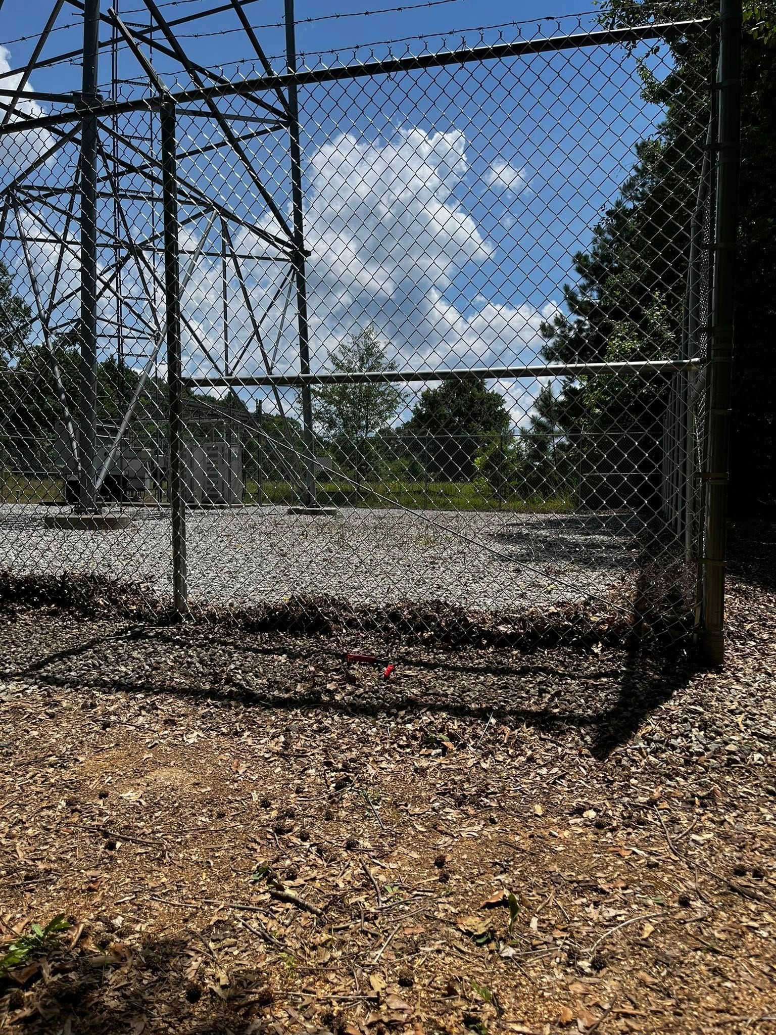 Chain link fence encloses a structure, with a blue sky and trees in the background.