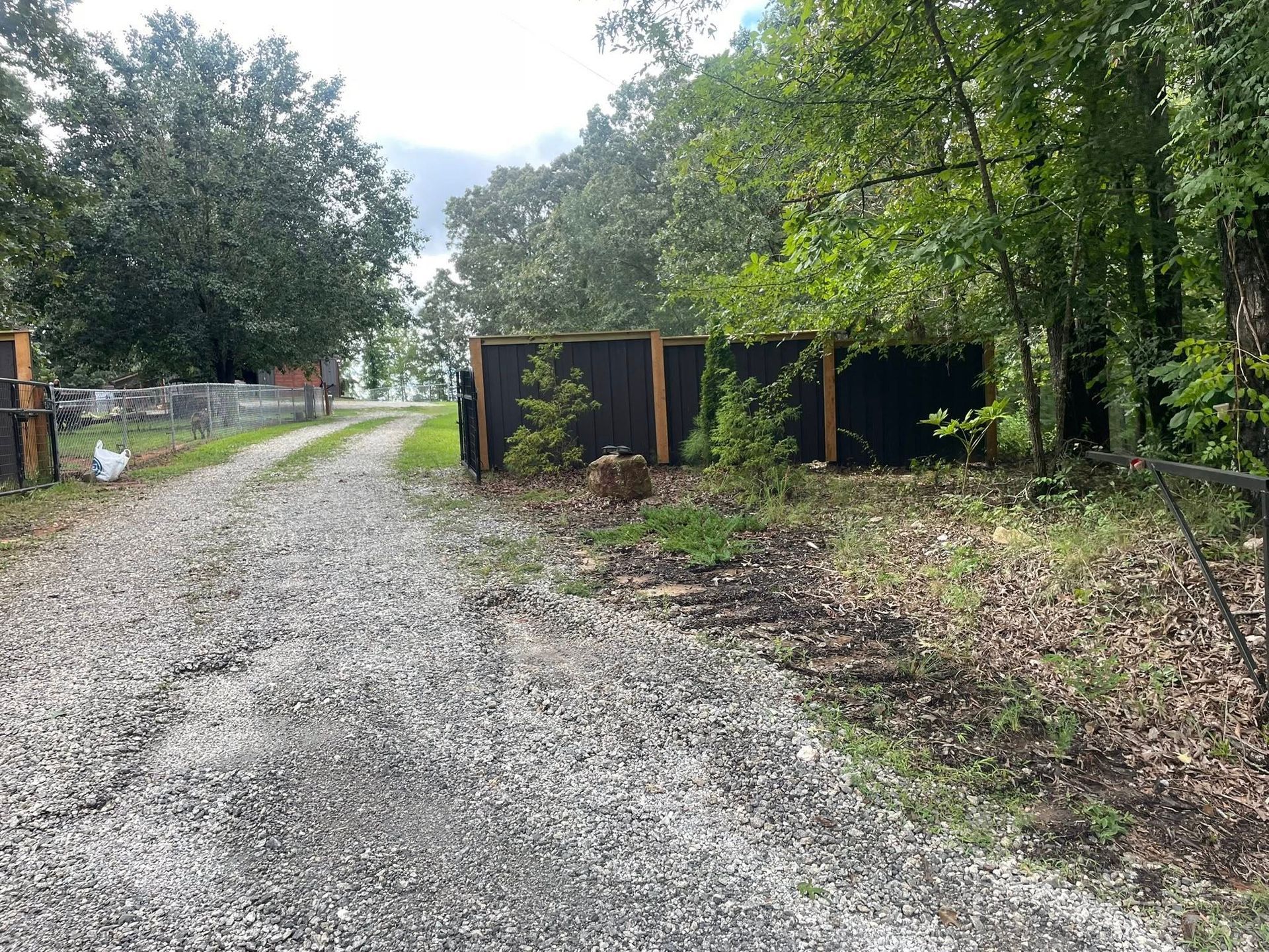 Gravel driveway leads to a fenced area with trees. Black fabric covers the wooden fence.