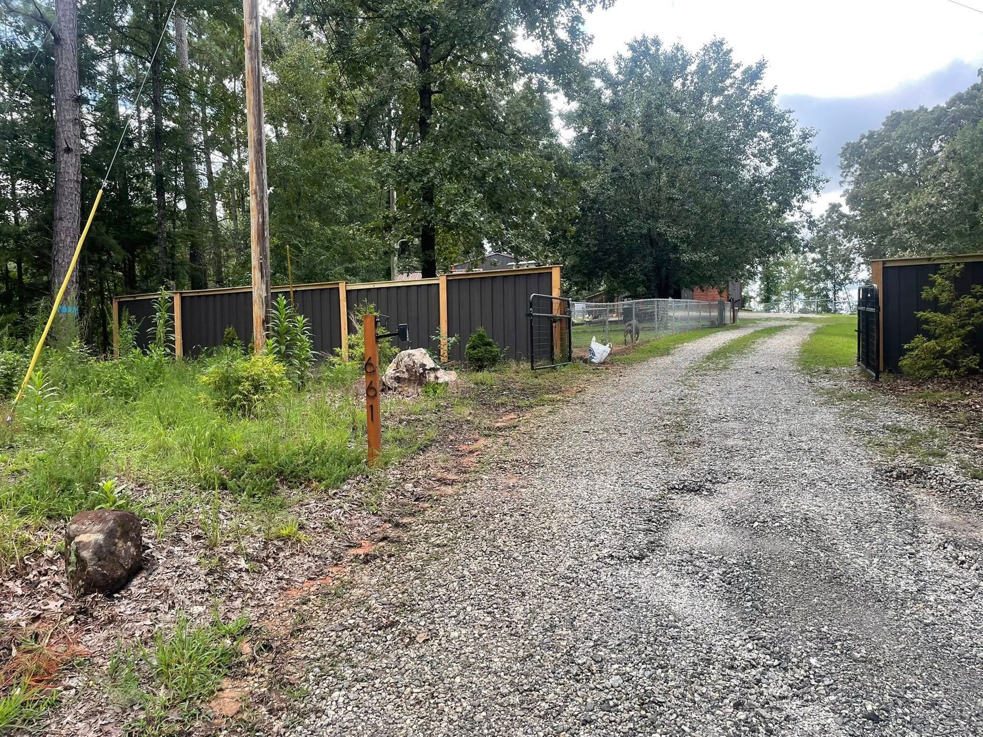 Gravel driveway leads to a property with a dark fence in a wooded area on a cloudy day.