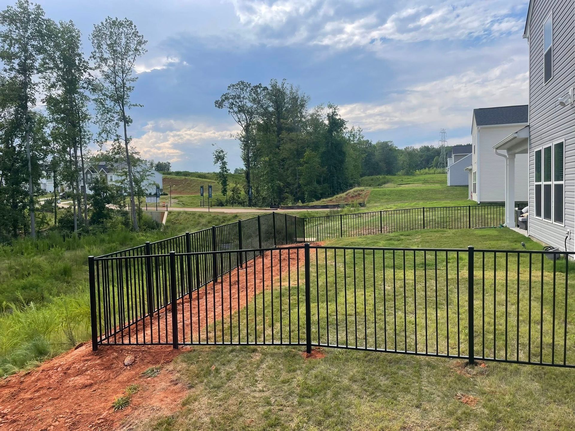 Black metal fence surrounds a grassy yard next to a residential building and a small dirt path under a cloudy sky.