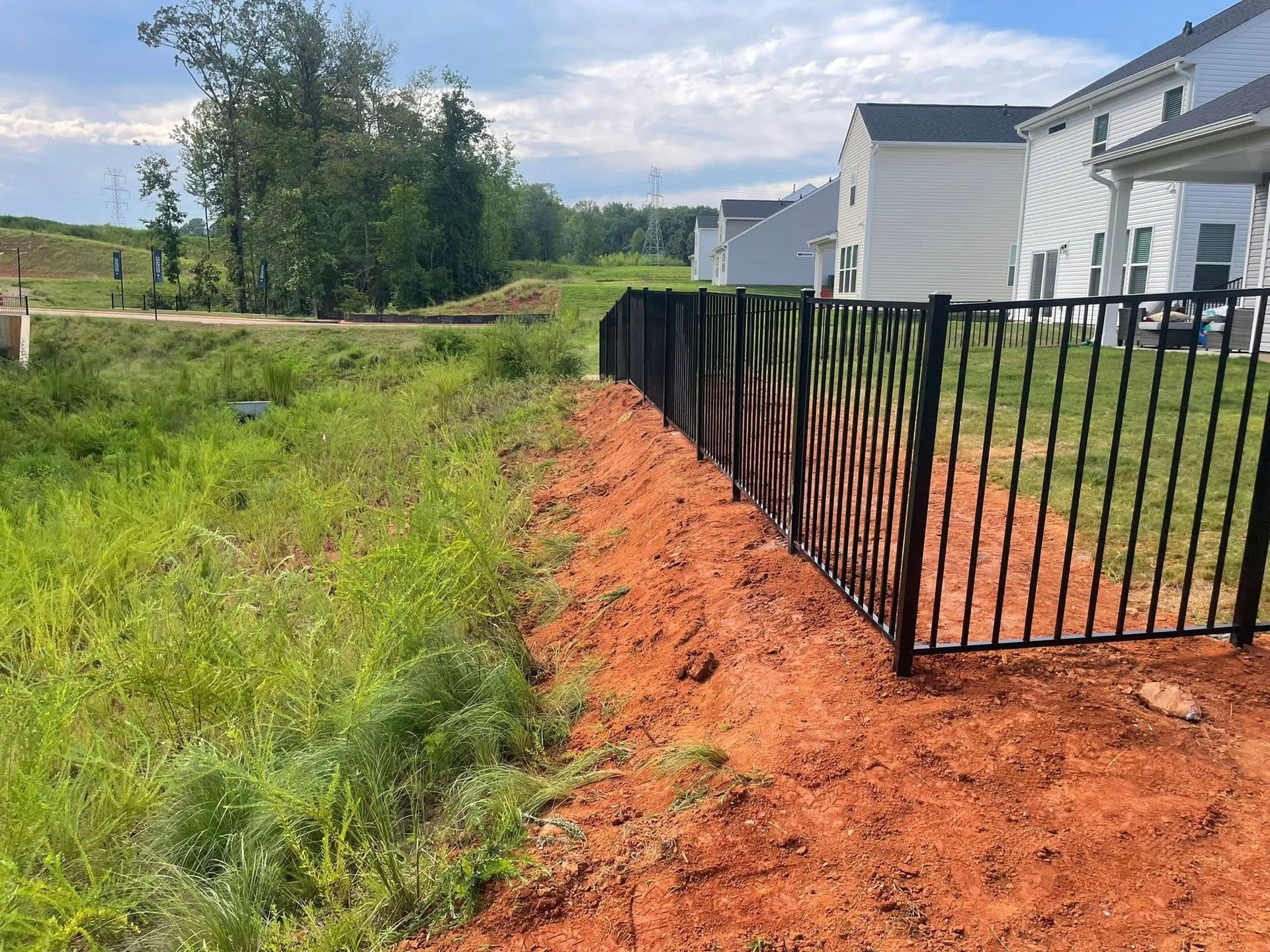 Black metal fence along red soil, separating tall green grass from houses under a blue sky.