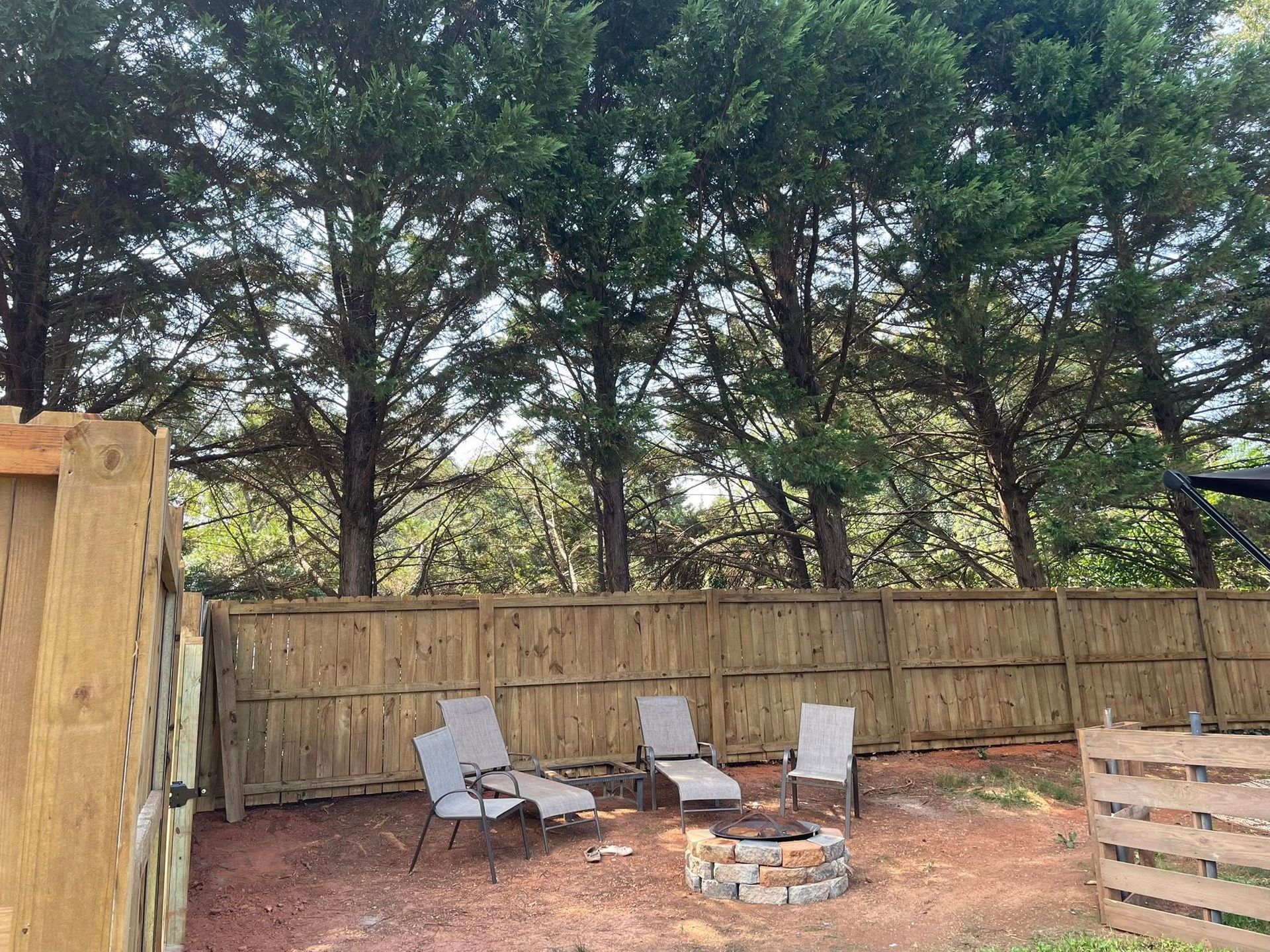 Backyard with a fire pit, chairs, and a wooden fence, trees in the background.