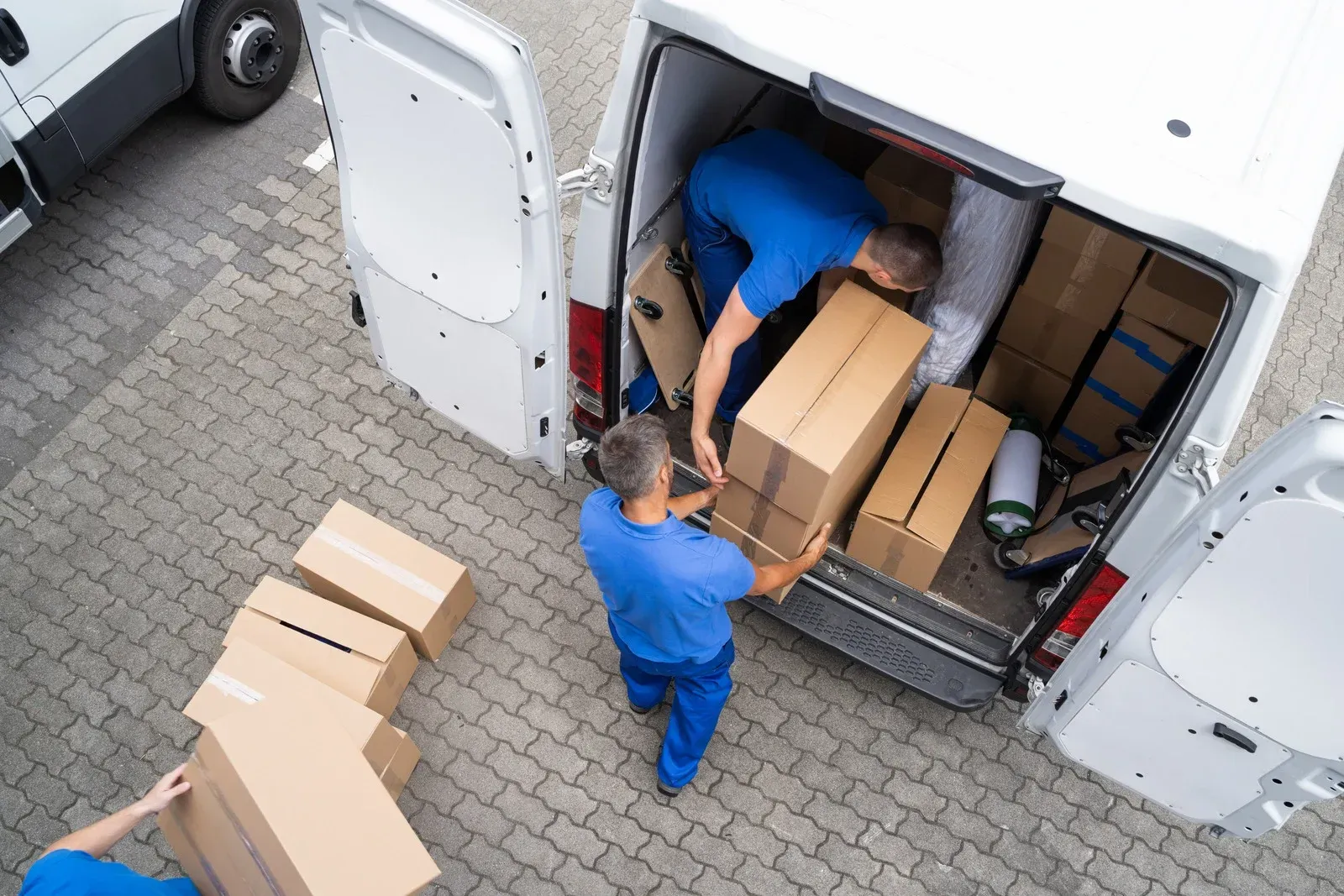 Movers in blue unloading boxes from a white van on a brick paved surface.