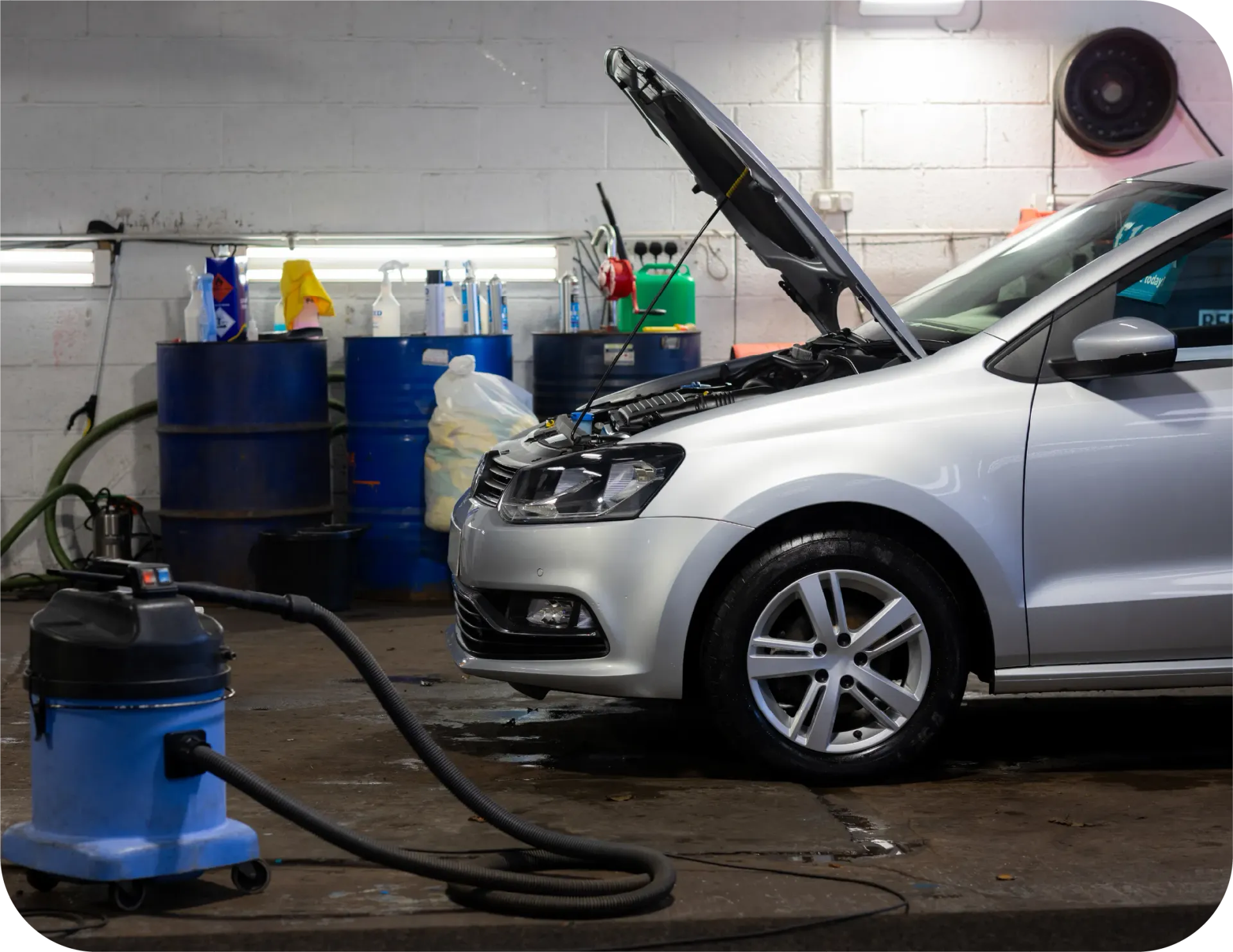 A silver car with its hood open is being cleaned by a vacuum cleaner.