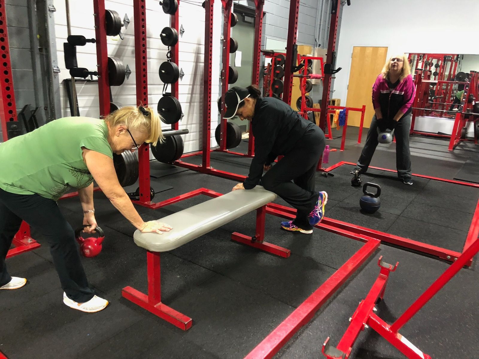 Three women exercise with kettlebells in a red-themed gym. One woman leans on a bench, another hoists a kettlebell, and a third is about to swing a weight.