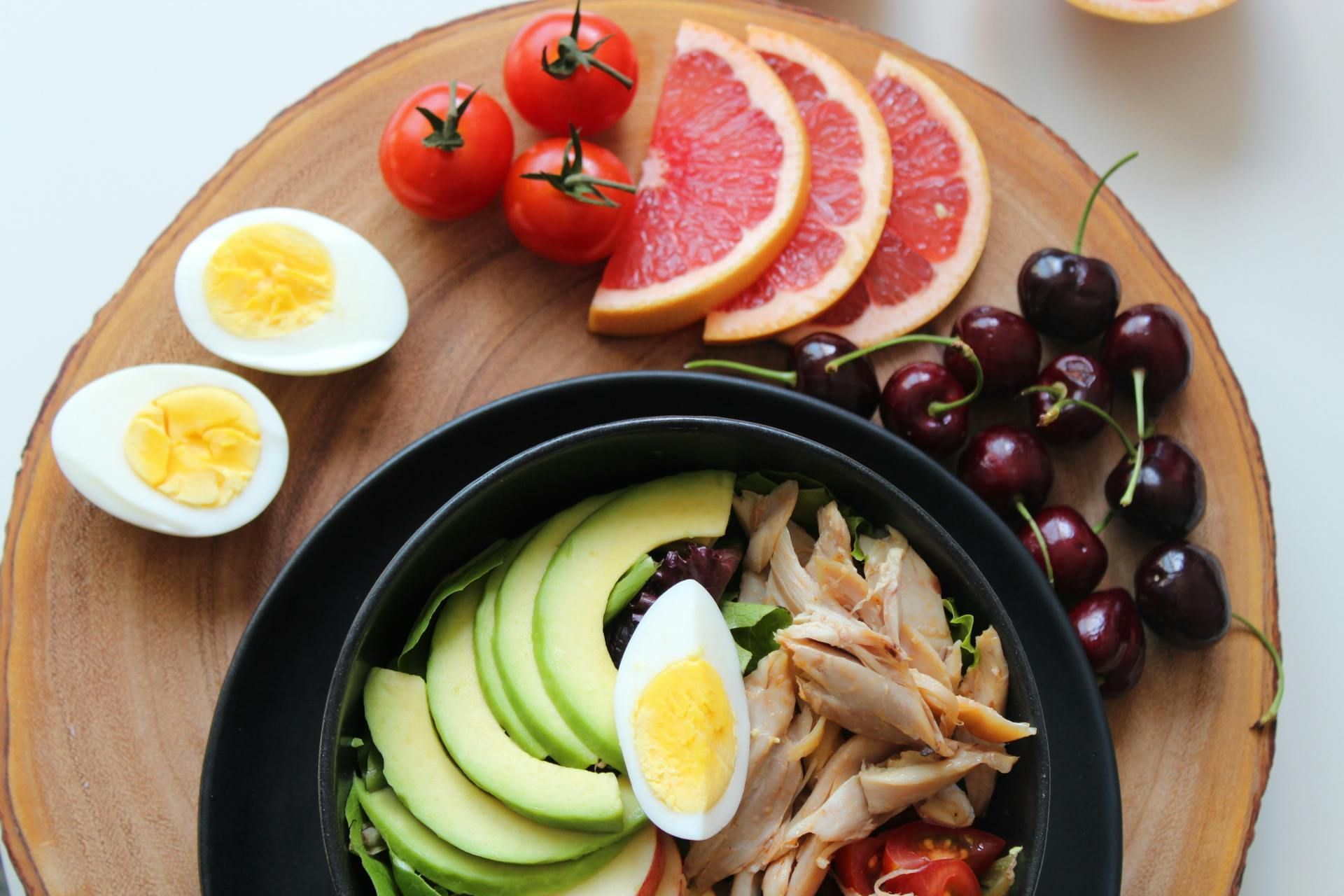 Healthy breakfast bowl with avocado, eggs, and chicken, alongside grapefruit slices, cherries, and tomatoes on a wooden board.