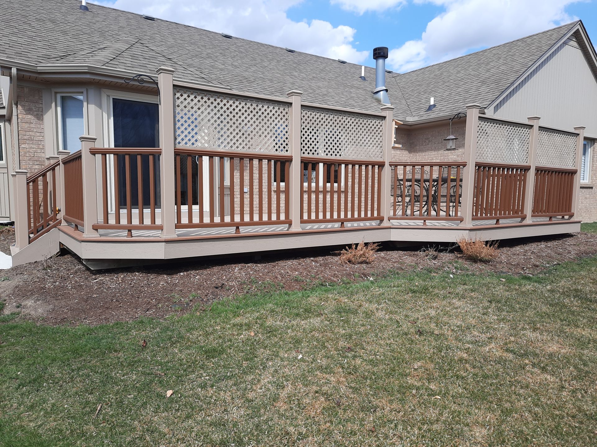 Brown wooden deck attached to a beige house, with lattice and railing, on a grassy lawn.