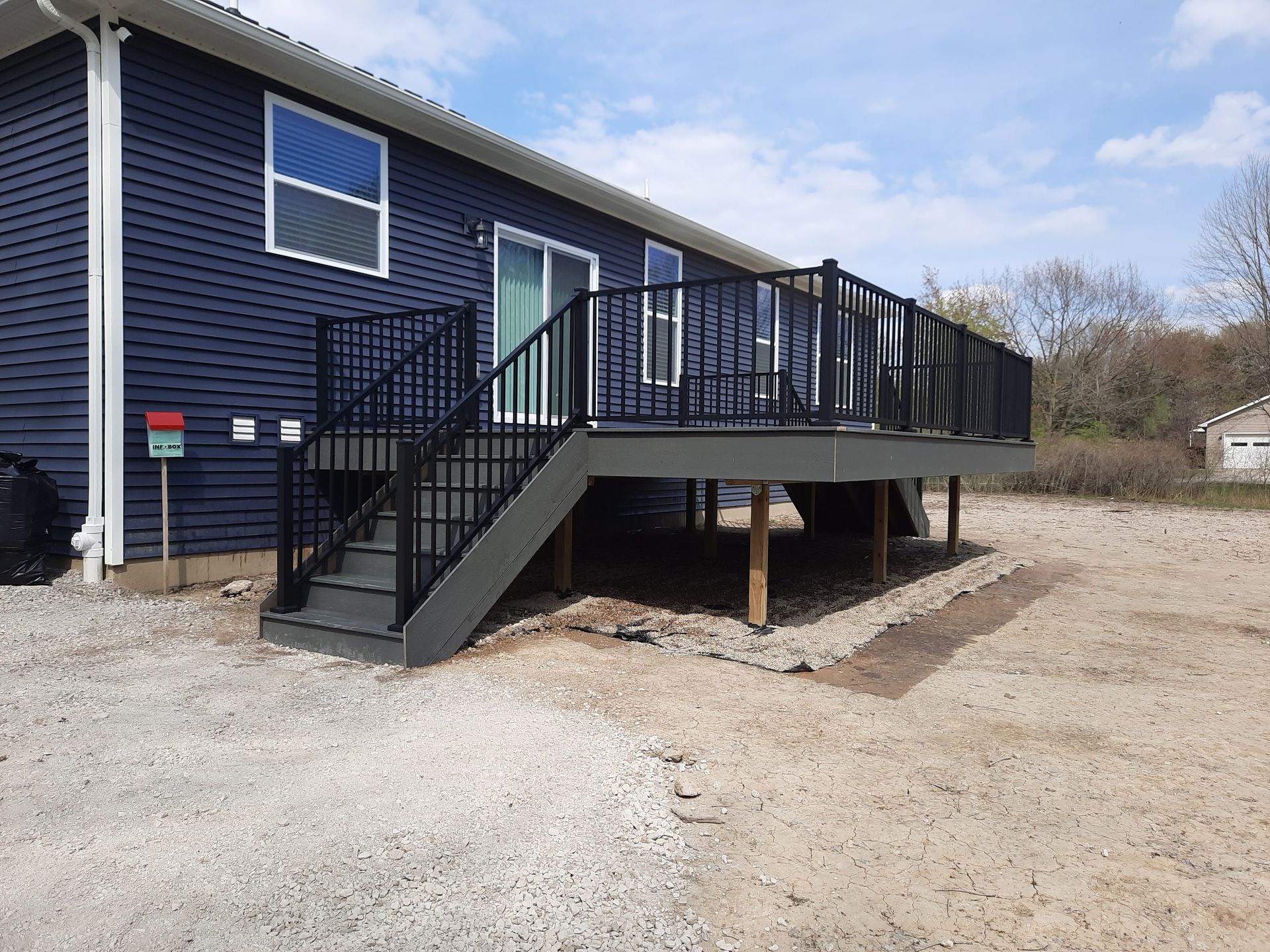 A blue house with a gray deck and black railing. Gravel ground and a cloudy sky.