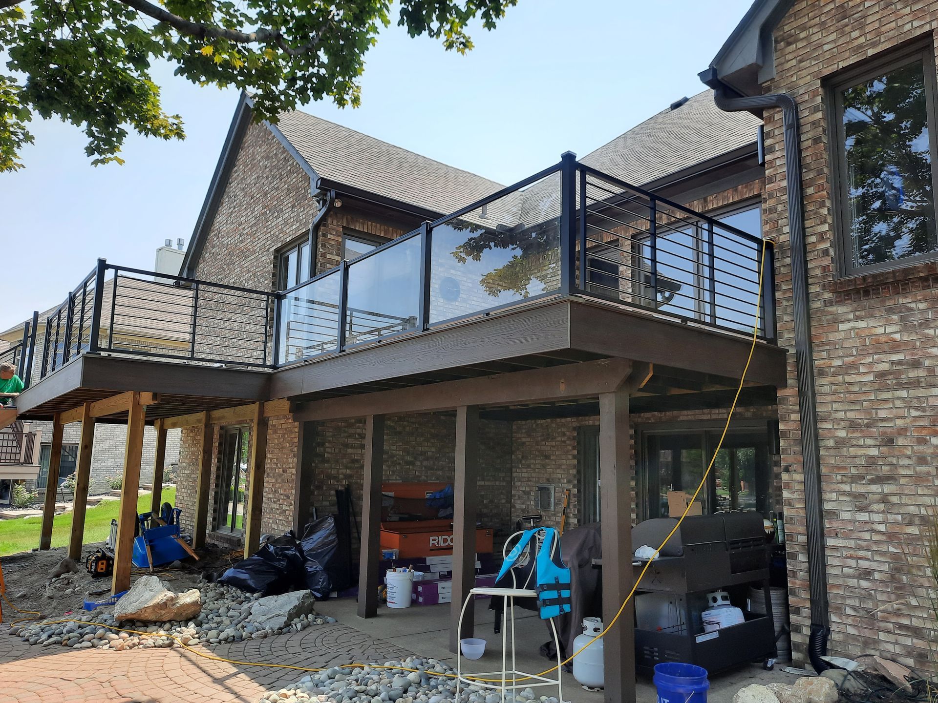 Two-story brick house with a deck featuring glass and black metal railing. Lower level has patio furniture and a grill.