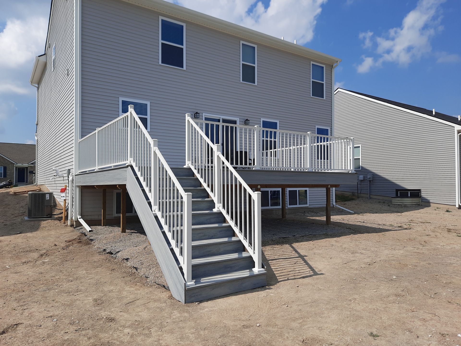 Back of a two-story gray house with a white-railed deck and stairs.  Setting is a yard with dirt and a clear sky.