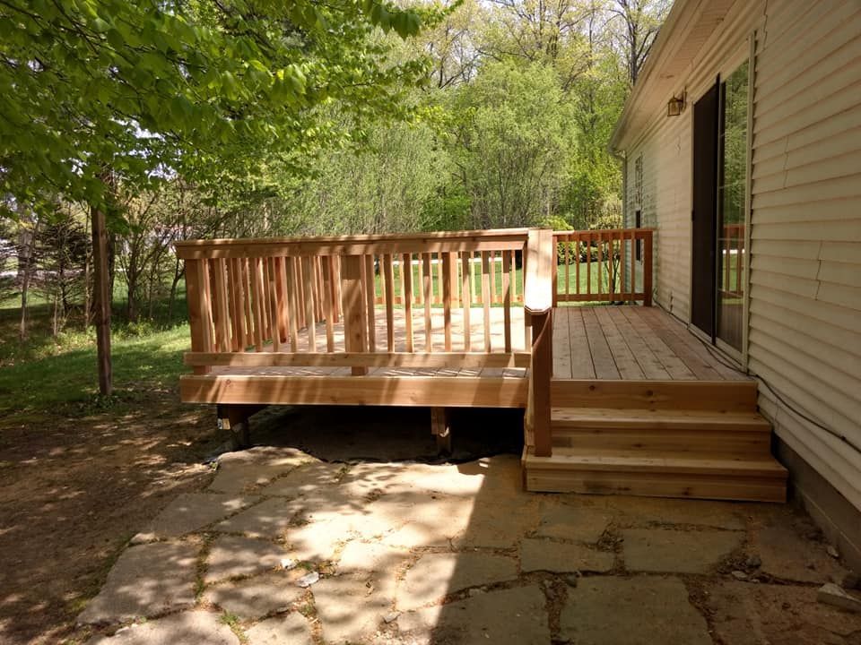 Wooden deck with steps leading to a stone patio next to a house with a sliding glass door, surrounded by trees.