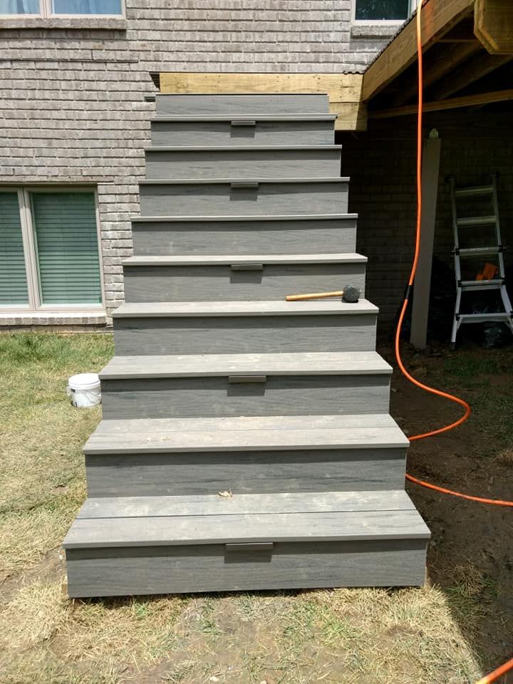 Gray outdoor stairs leading up to a deck. A hammer rests on a step. Brick building and ladder in the background.