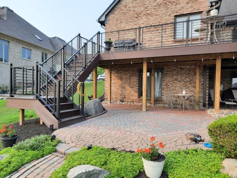 Brick patio with deck and stairs, metal railings. Brown deck, brick house, green yard, potted flowers.