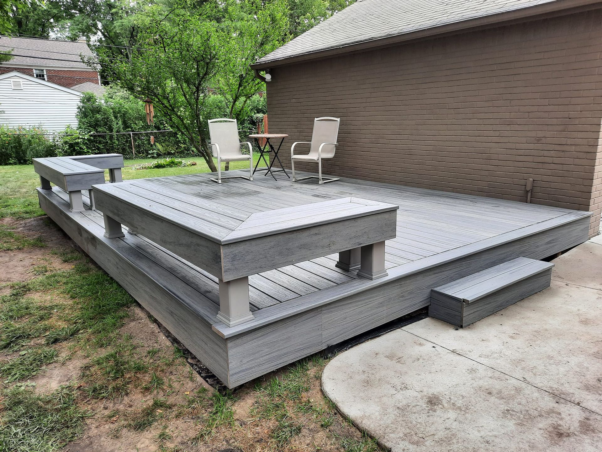 Gray composite deck with built-in benches, two chairs, and small table near a house with greenery in the background.