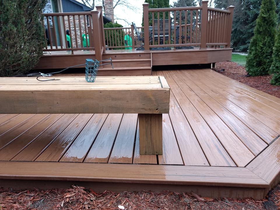 Brown wooden deck with built-in bench, steps, and railing. Wet surface, surrounded by mulch and greenery.