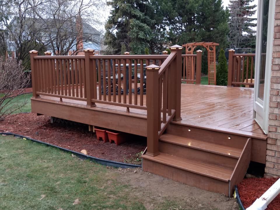 Brown deck with steps, railings, and arbor in a backyard. Wet deck boards, grass and mulch border.