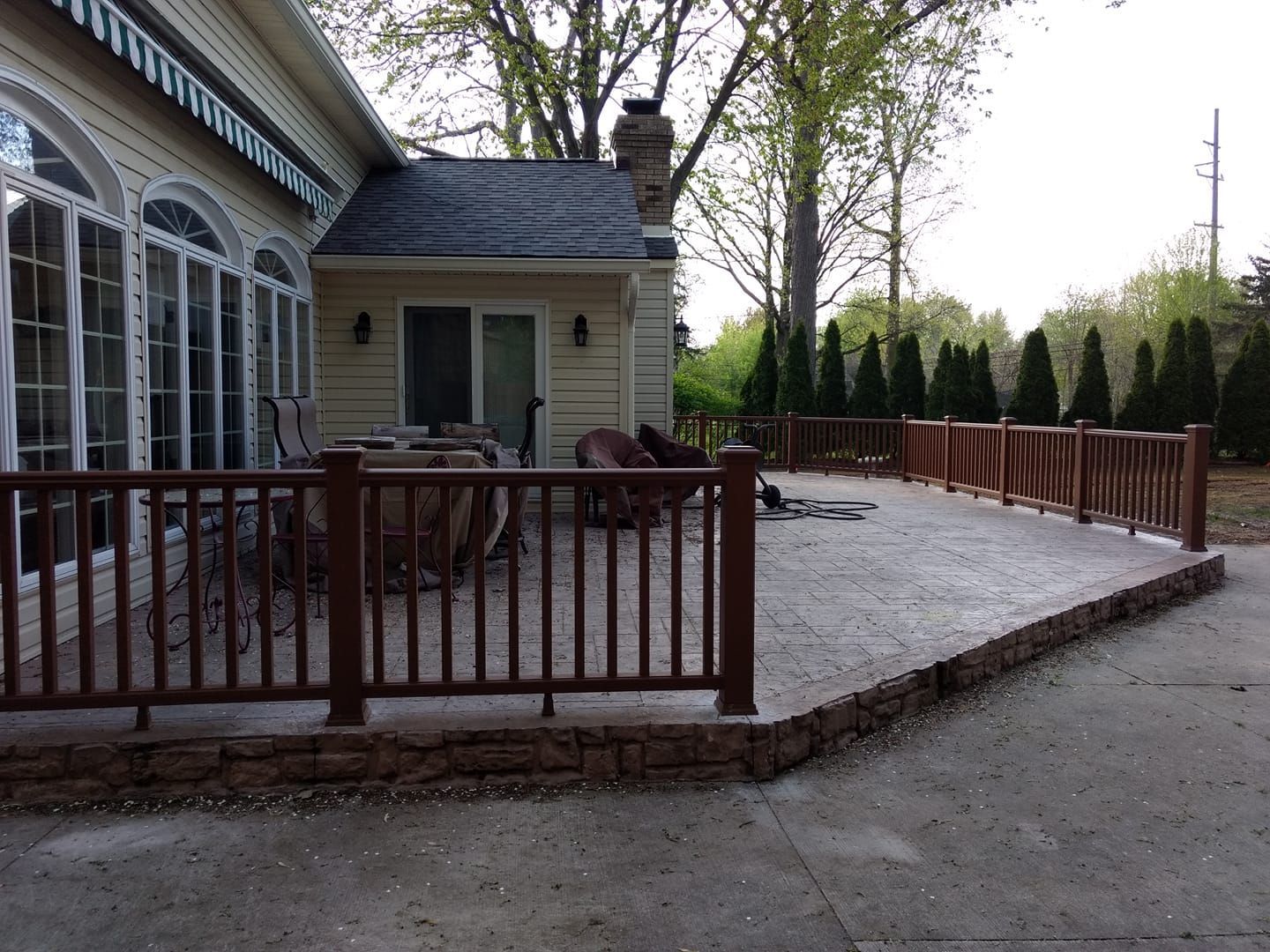 Brown wooden deck and railing with concrete patio, next to a beige house with large windows.