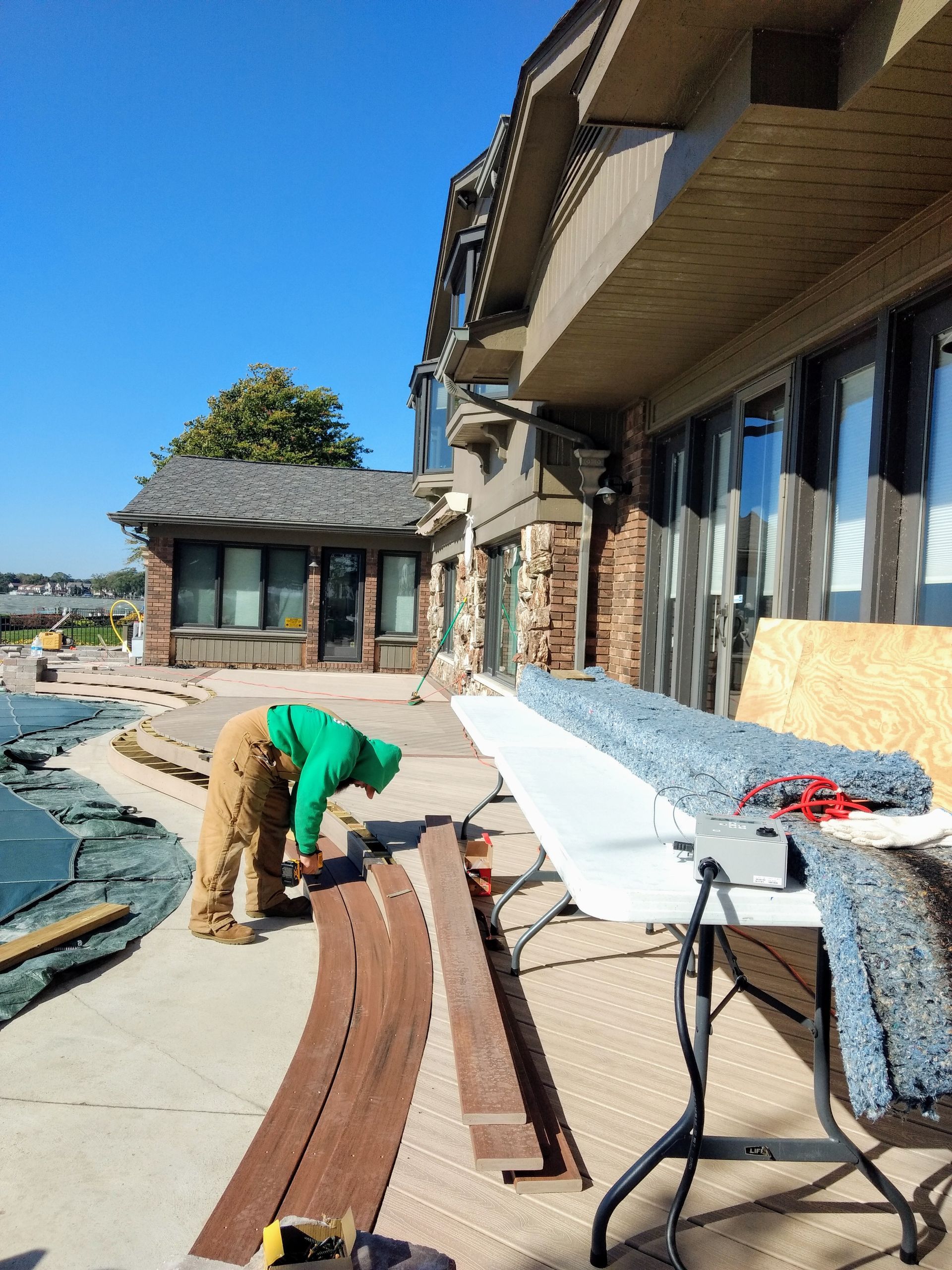 A person works on a curved wooden structure outdoors, with a building and table in the background.