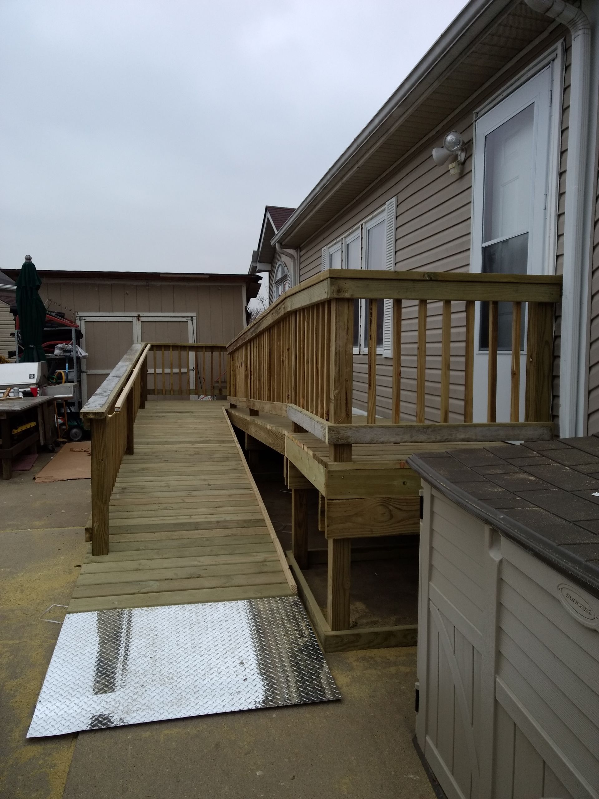 Wooden ramp leading to a deck beside a house with tan siding and a shed.