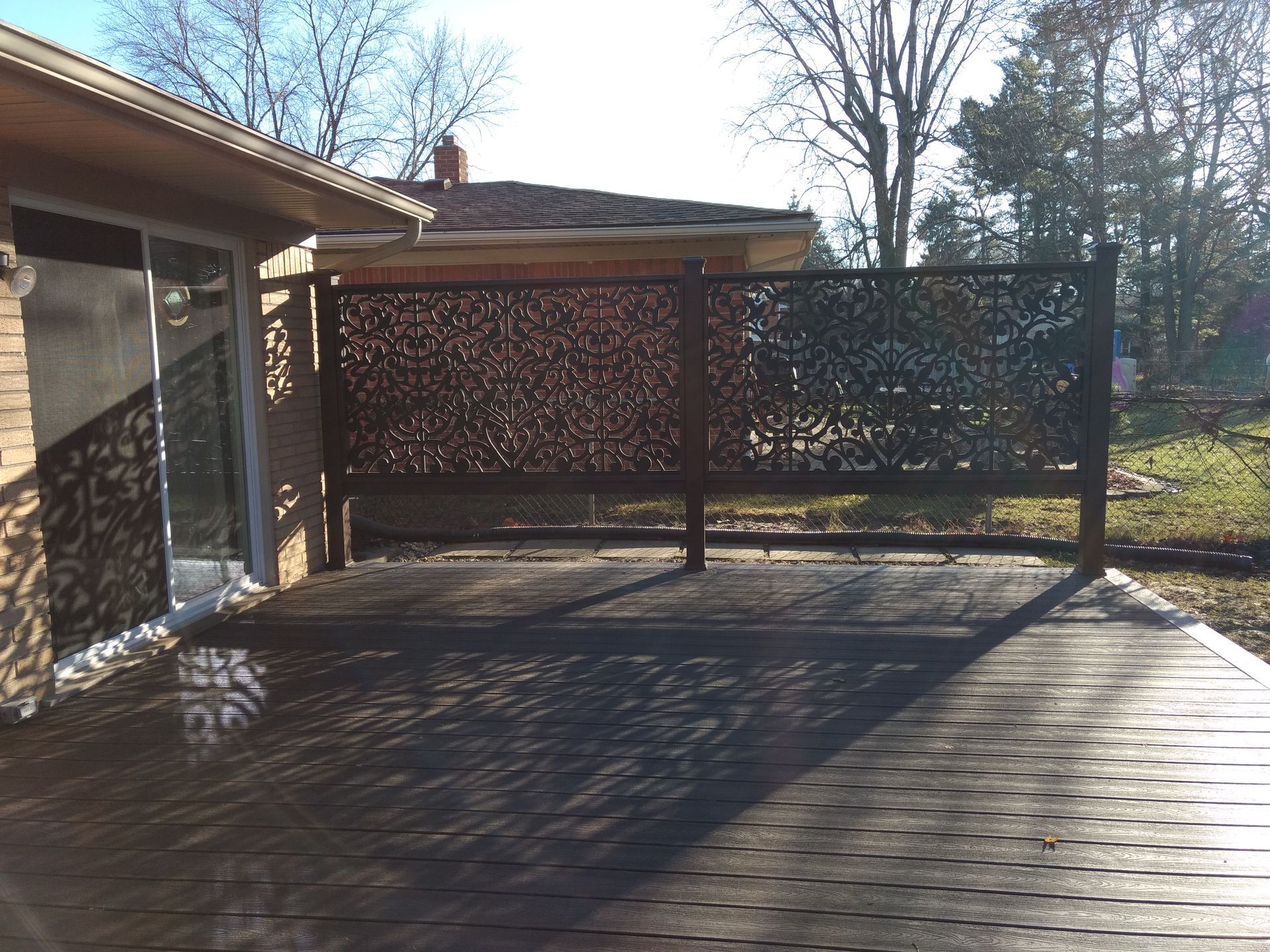 A wooden deck with a decorative metal screen against a house and yard. Sunny day.