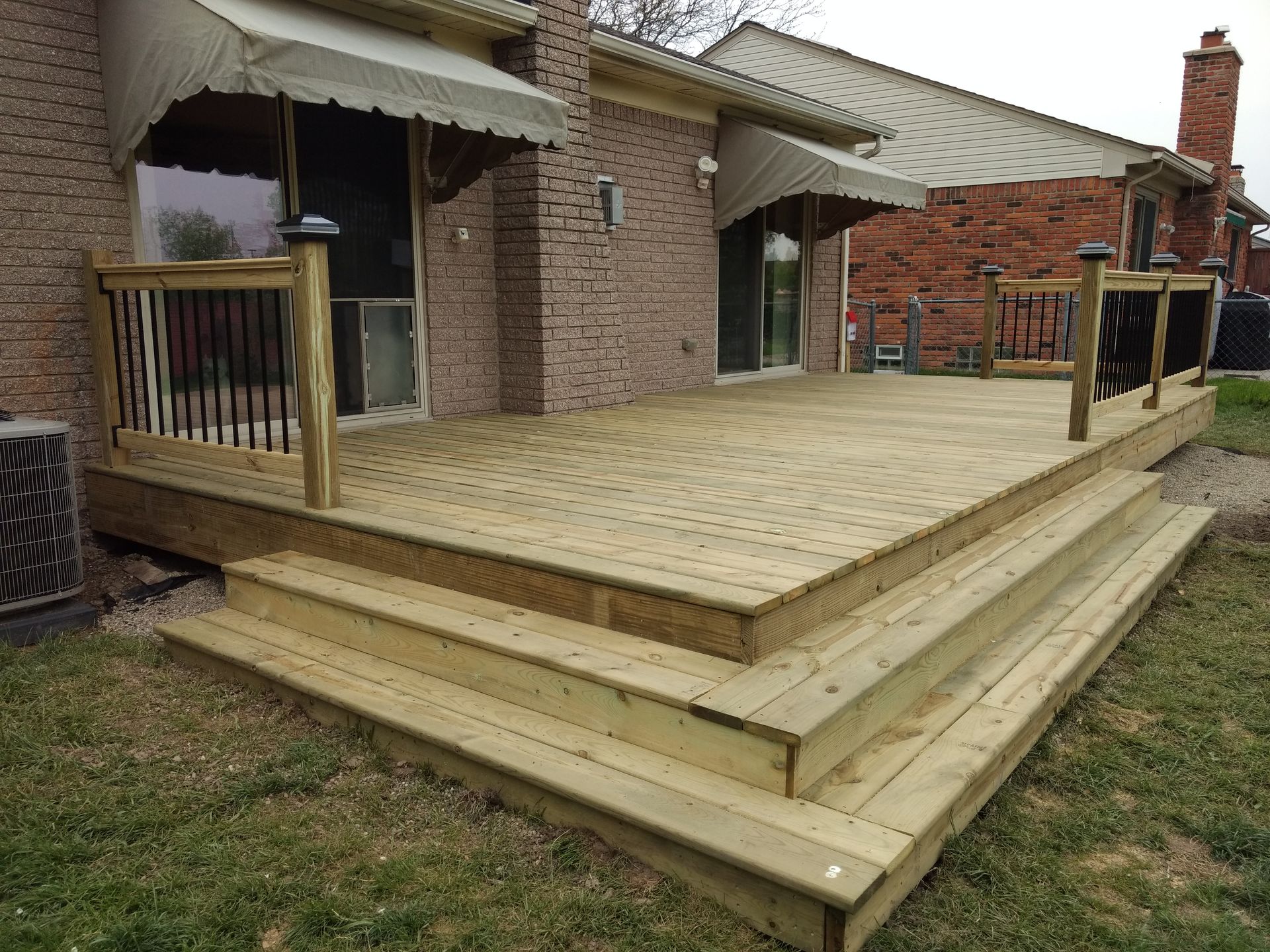 Wooden deck with steps attached to a brick house, featuring black railings and awnings.